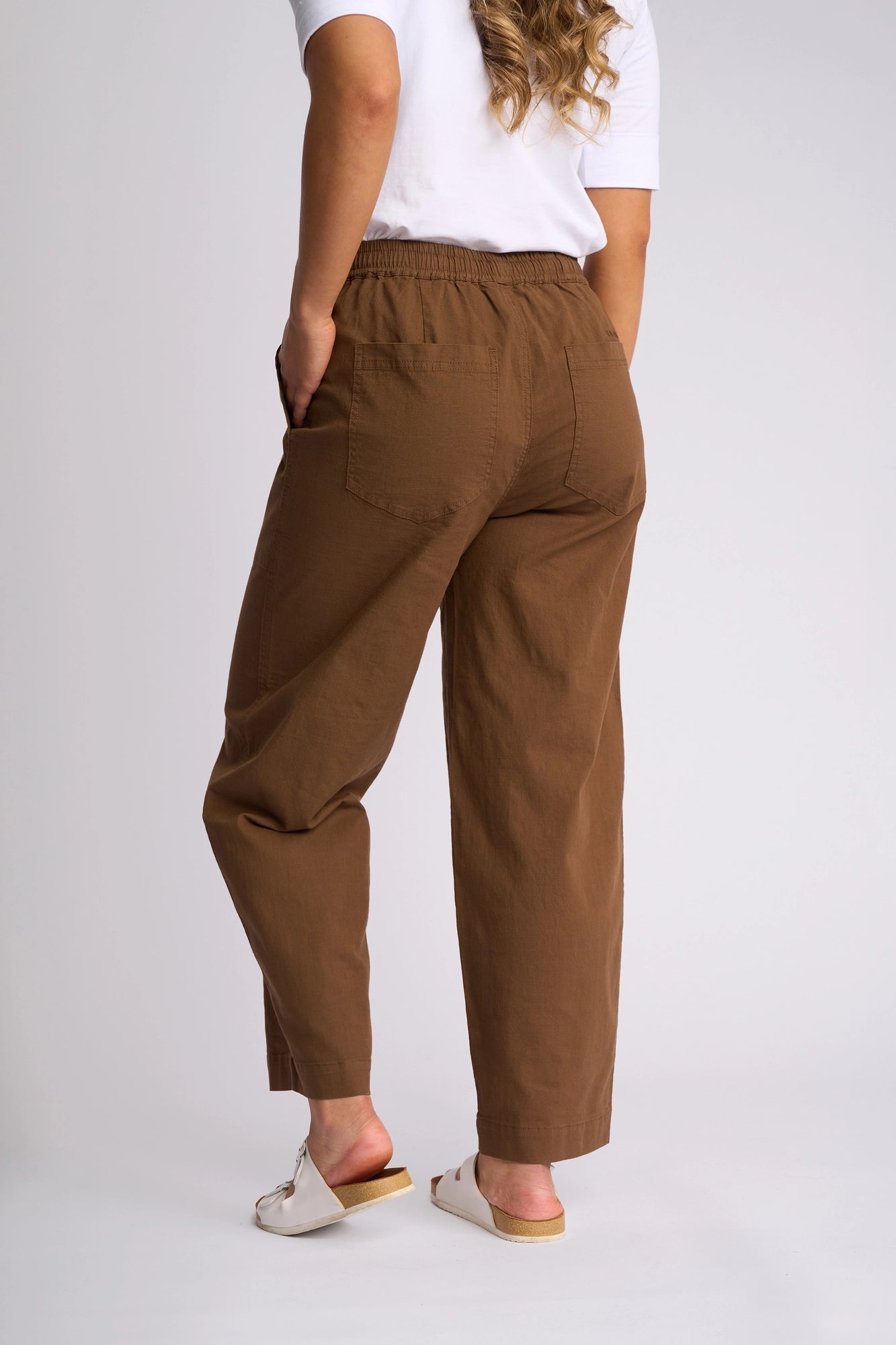 A person wearing a white shirt, the Vassalli Dione Pant in Pecan—crafted from stretch cotton with an elasticated drawstring waistband—and white slide sandals, stands with hands in pockets facing away from the camera.