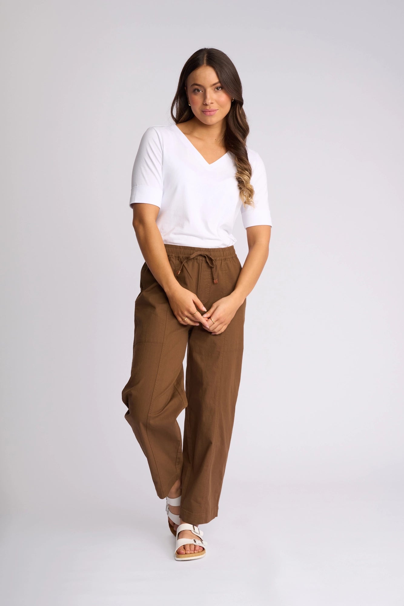 A woman stands against a light background wearing a white V-neck shirt, the Vassalli Dione Pant in Pecan featuring a loose barrel-leg silhouette and an elasticated drawstring waistband, and white sandals. Her long hair is styled in waves, she has a neutral expression, and her hands are loosely clasped in front.