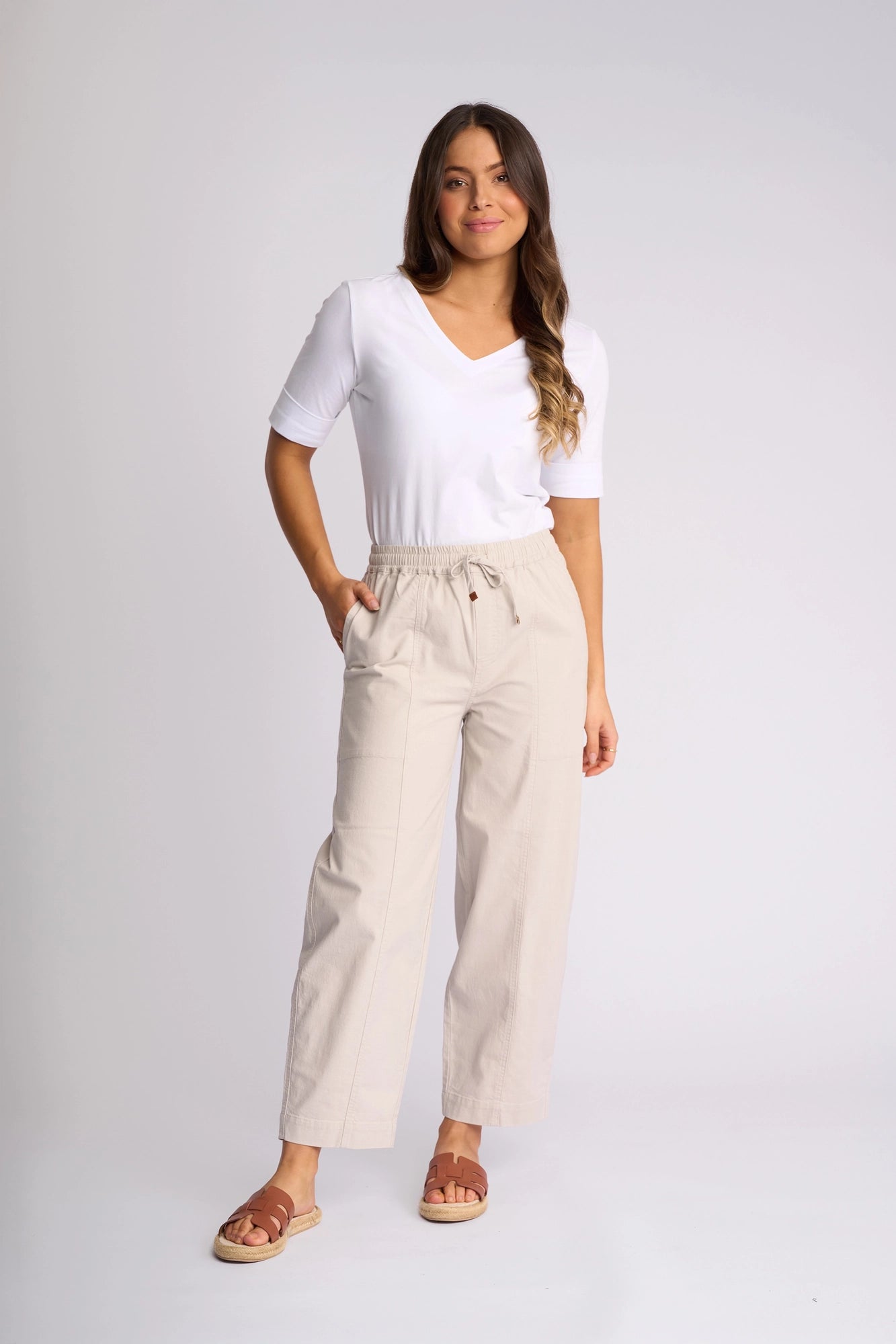 A woman stands against a plain background, wearing a white short-sleeve top paired with the Vassalli Dione Pant in Stone—beige barrel-leg pants featuring an elasticated waistband and pockets—and brown sandals. She has long, wavy hair and poses in a relaxed, casual manner with a slight smile.