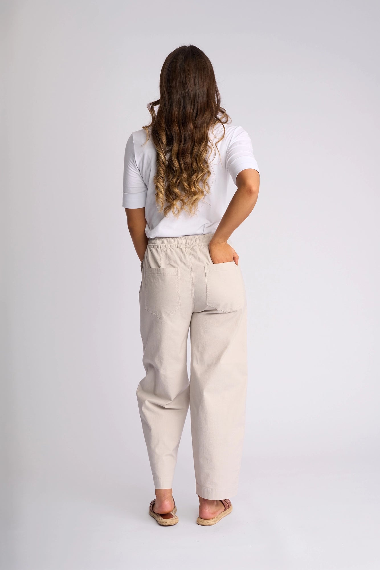 A woman with long wavy hair, dressed in a white short-sleeve shirt, Vassalli’s Dione Pant - Stone (a light beige stretch cotton barrel-leg pant with an elasticated waistband), and sandals, stands facing away against a plain light background with one hand in her pocket.