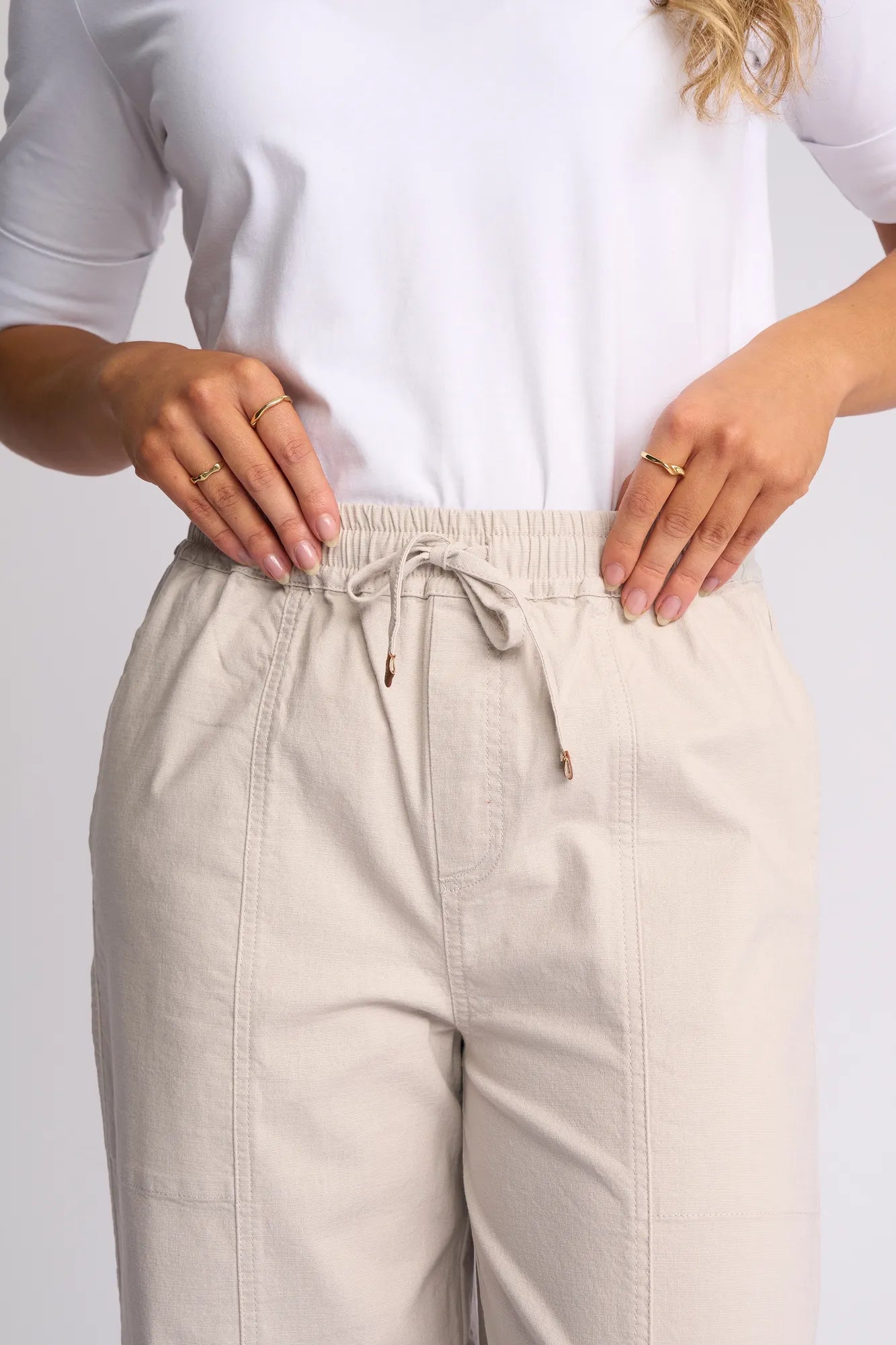 A person stands with hands on their hips, wearing the Vassalli Dione Pant in Stone—a light beige barrel-leg design with an elasticated waistband—and a white shirt, while several gold rings adorn their fingers.