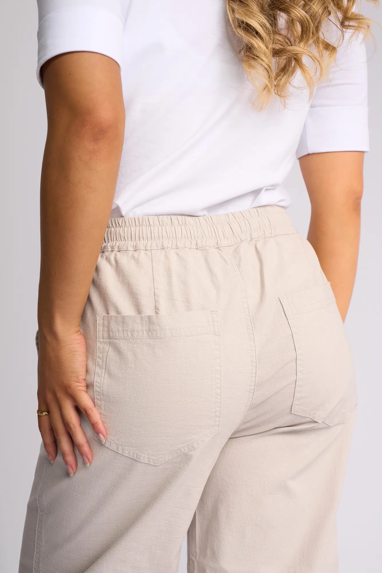 A person wearing a white shirt and the Vassalli Dione Pant in Stone—featuring a barrel-leg silhouette with an elasticated waistband—is shown from the back, one hand resting on their hip and their long, wavy hair partially visible.