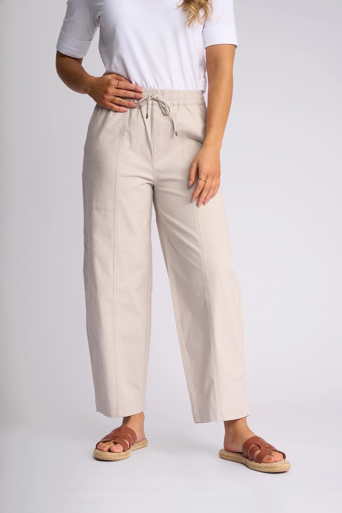 A woman poses with one hand on her waist against a plain white background, wearing Vassalli’s Dione Pant in Stone—a beige barrel-leg style with an elasticated waistband—paired with a white short-sleeve top and tan sandals. Her face is not visible.