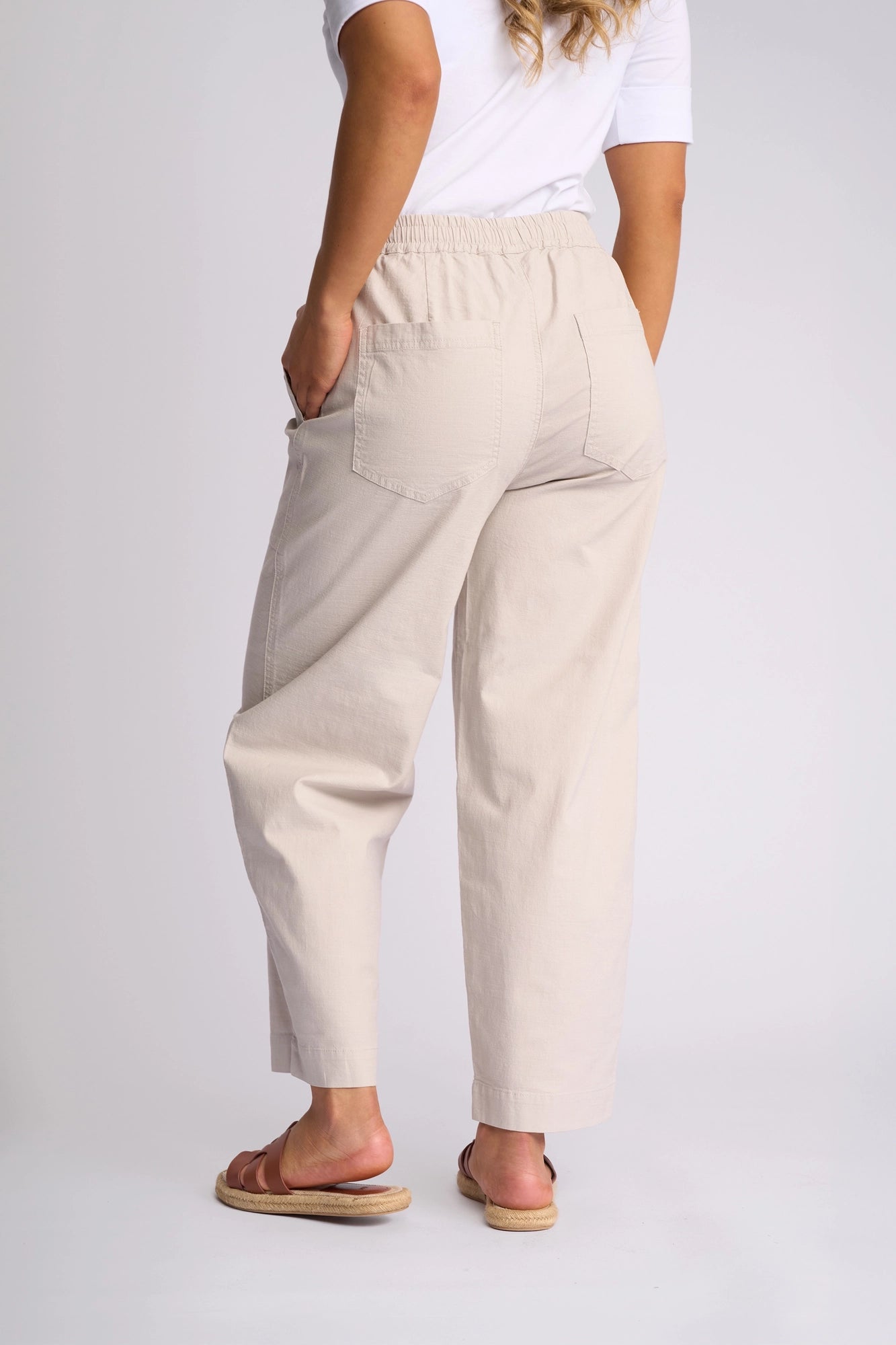 A person is standing barefoot and facing away, wearing the Vassalli Dione Pant in Stone—a light beige, loose-fitting barrel-leg style made from stretch cotton. These pants feature an elasticated waistband and two back pockets, combining comfort with relaxed style.
