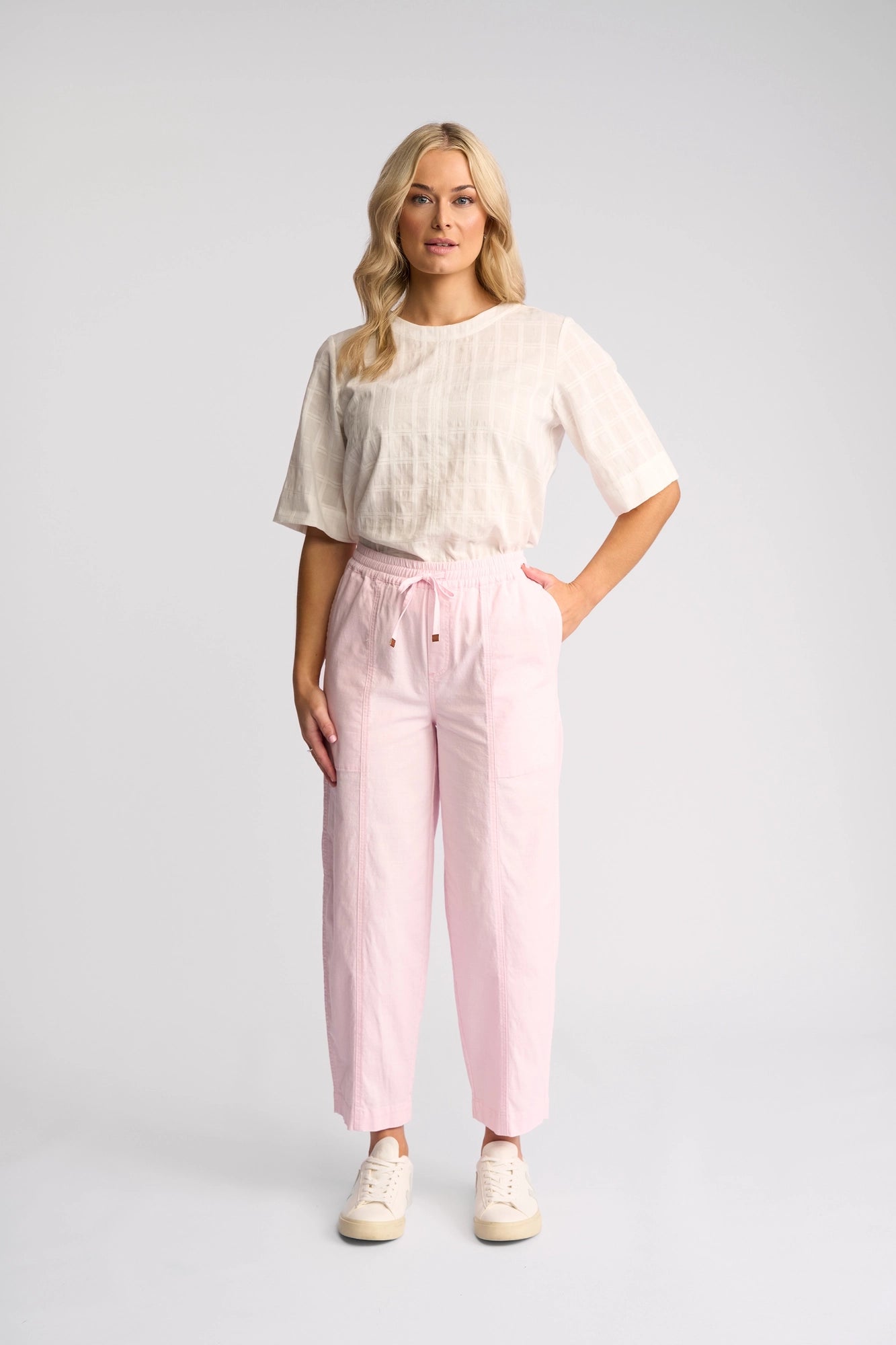 A woman stands facing forward against a plain light background, wearing a loose white short-sleeve top, the Vassalli Dione Pant in Taffy—light pink stretch cotton with a barrel-leg, ankle-grazer silhouette—and white sneakers.