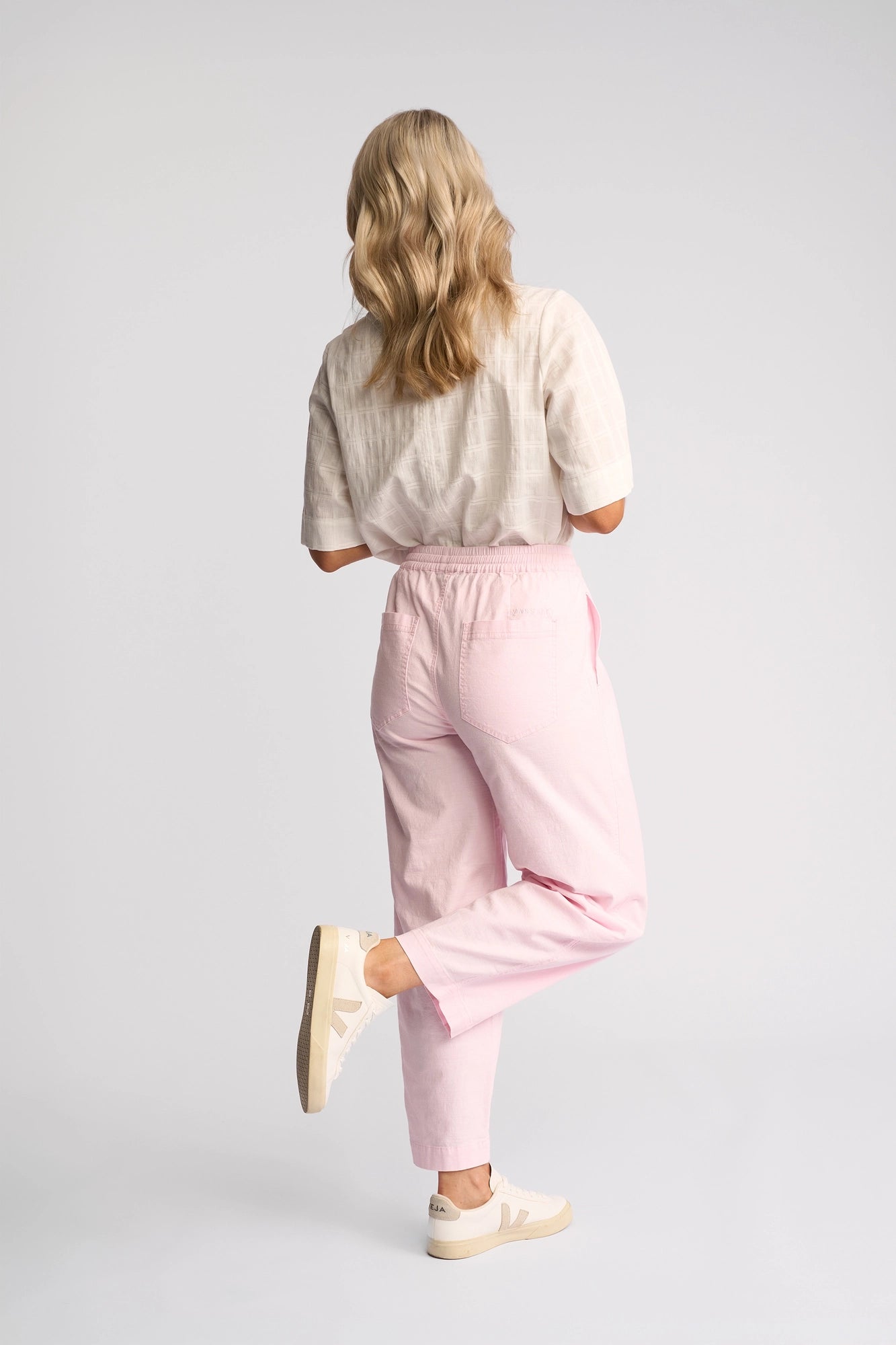 A person with wavy blonde hair, wearing a white shirt, Vassalli’s Dione Pant in Taffy—a light pink barrel-leg pant in soft stretch cotton—and white sneakers, stands on one leg with their back facing the camera against a plain light background.