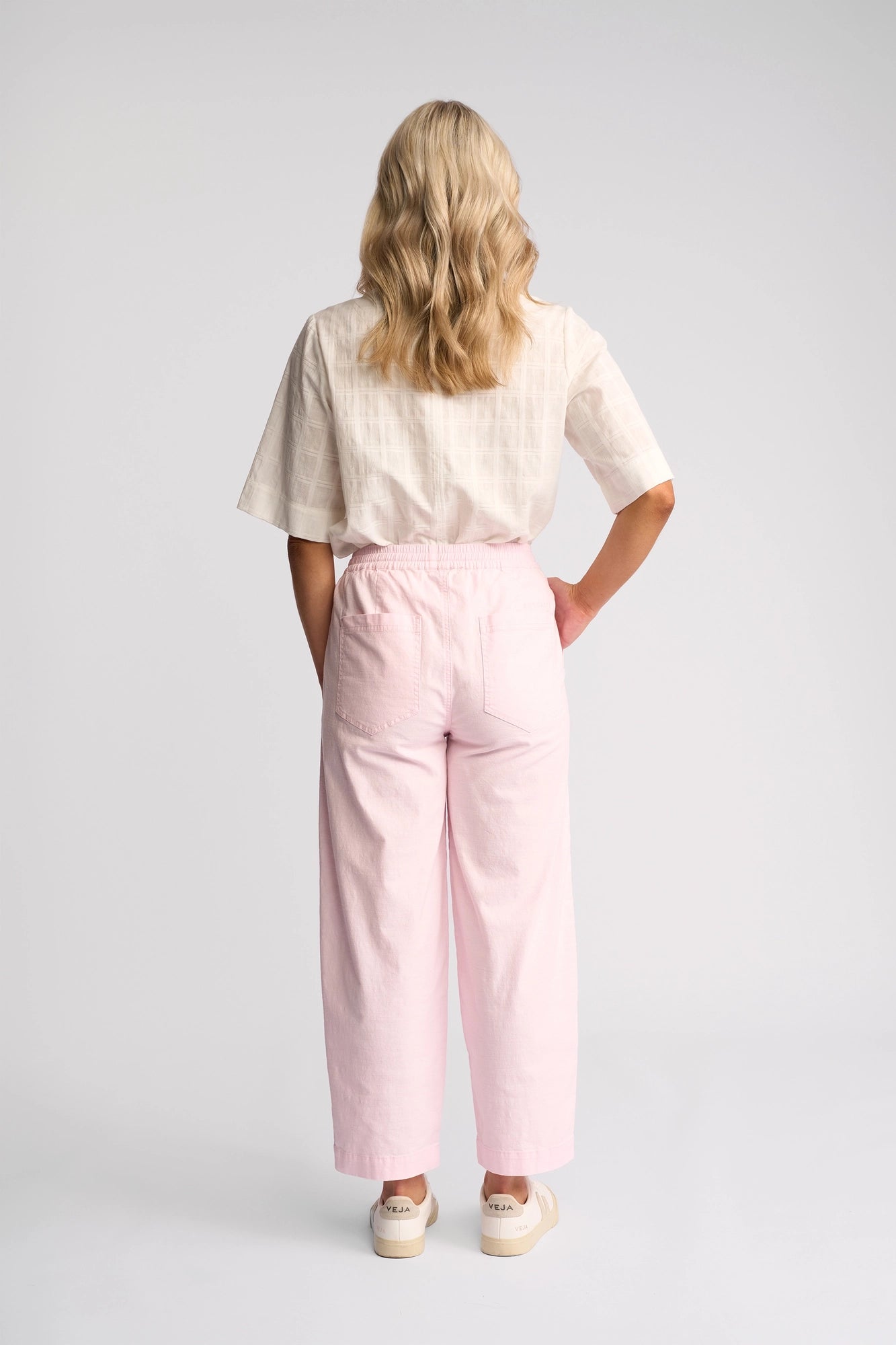 A person with wavy blonde hair stands facing away, wearing a light short-sleeve shirt, white sneakers, and the Vassalli Dione Pant - Taffy—high-waisted ankle-grazer pink barrel-leg pants made from stretch cotton—against a plain light background.