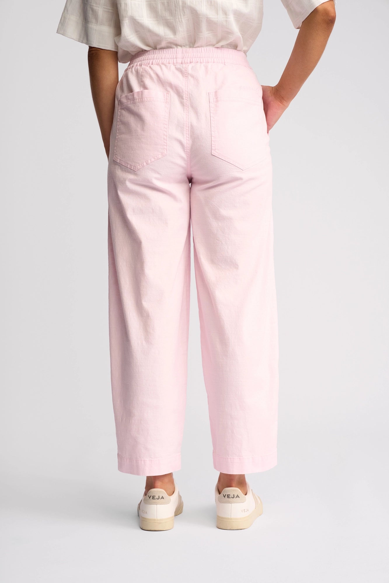 A person stands with their back to the camera, wearing the Vassalli Dione Pant in Taffy—a light pink, high-waisted ankle-grazer made from stretch cotton—paired with a white short-sleeve top and off-white Veja sneakers. The background is plain and light-colored.