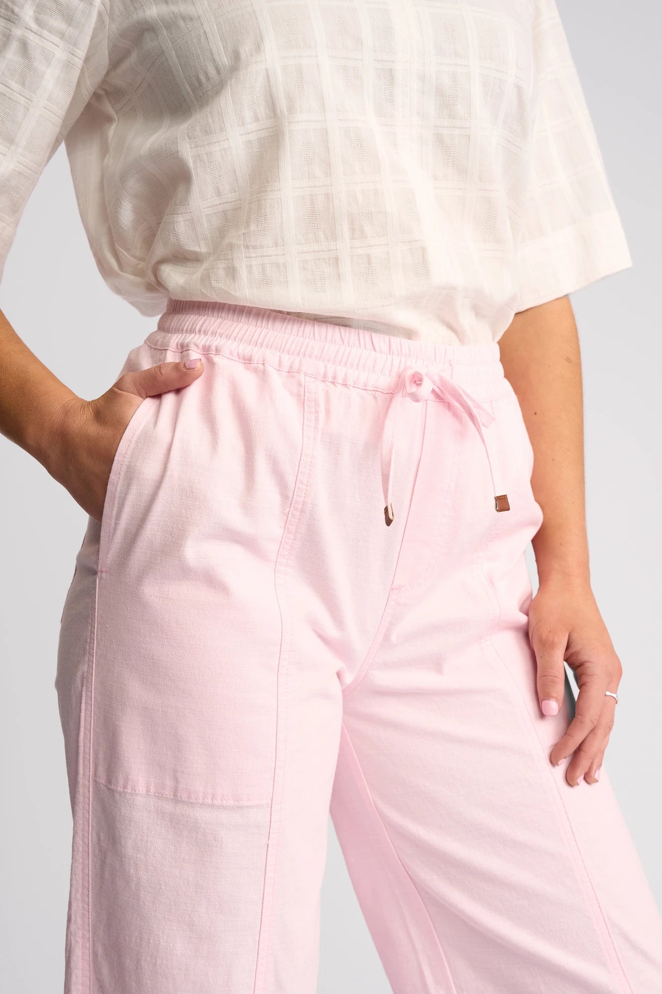 A woman wearing a white textured short-sleeve top and the Vassalli Dione Pant - Taffy, a light pink stretch cotton barrel-leg pant with pockets, stands with one hand in her pocket.