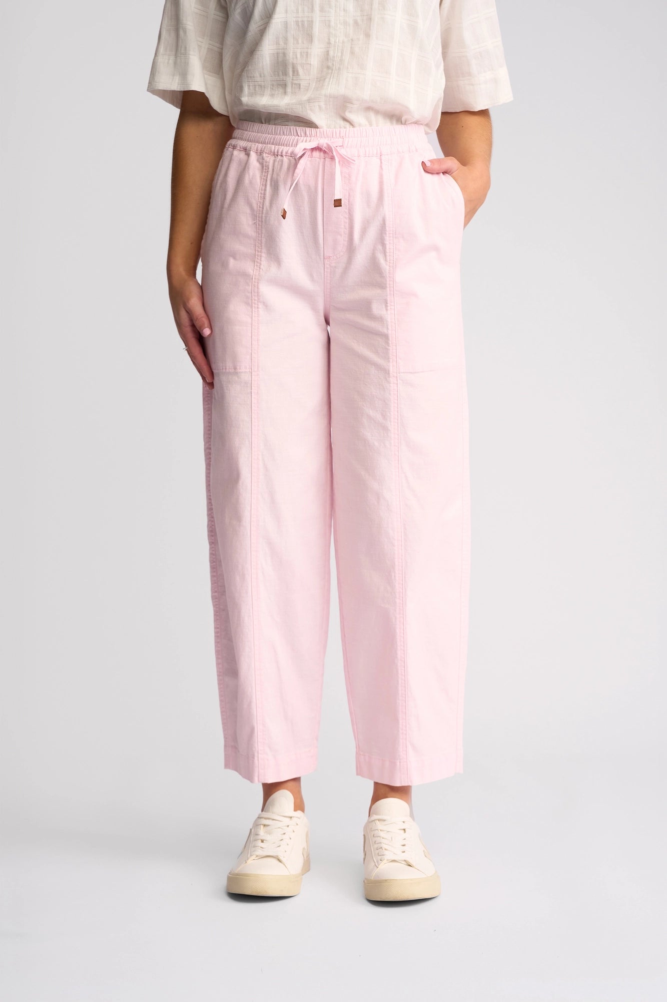 A person stands against a plain background wearing the Vassalli Dione Pant in Taffy—light pink, barrel-leg, ankle-grazer drawstring pants crafted from stretch cotton—paired with a light-patterned top and white sneakers. Their hands are in their pockets, and their face is not visible.