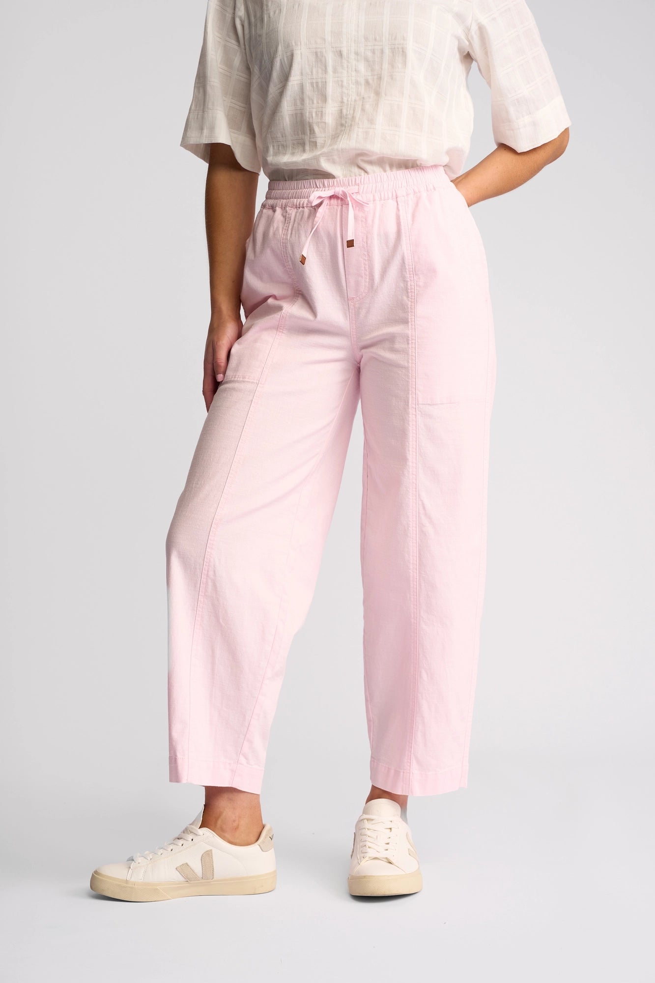 A person wearing a short-sleeve, light plaid top, the Vassalli Dione Pant in Taffy—a loose, light pink ankle-grazer barrel-leg style crafted from soft stretch cotton—and beige sneakers stands with one hand on their hip against a plain white background.