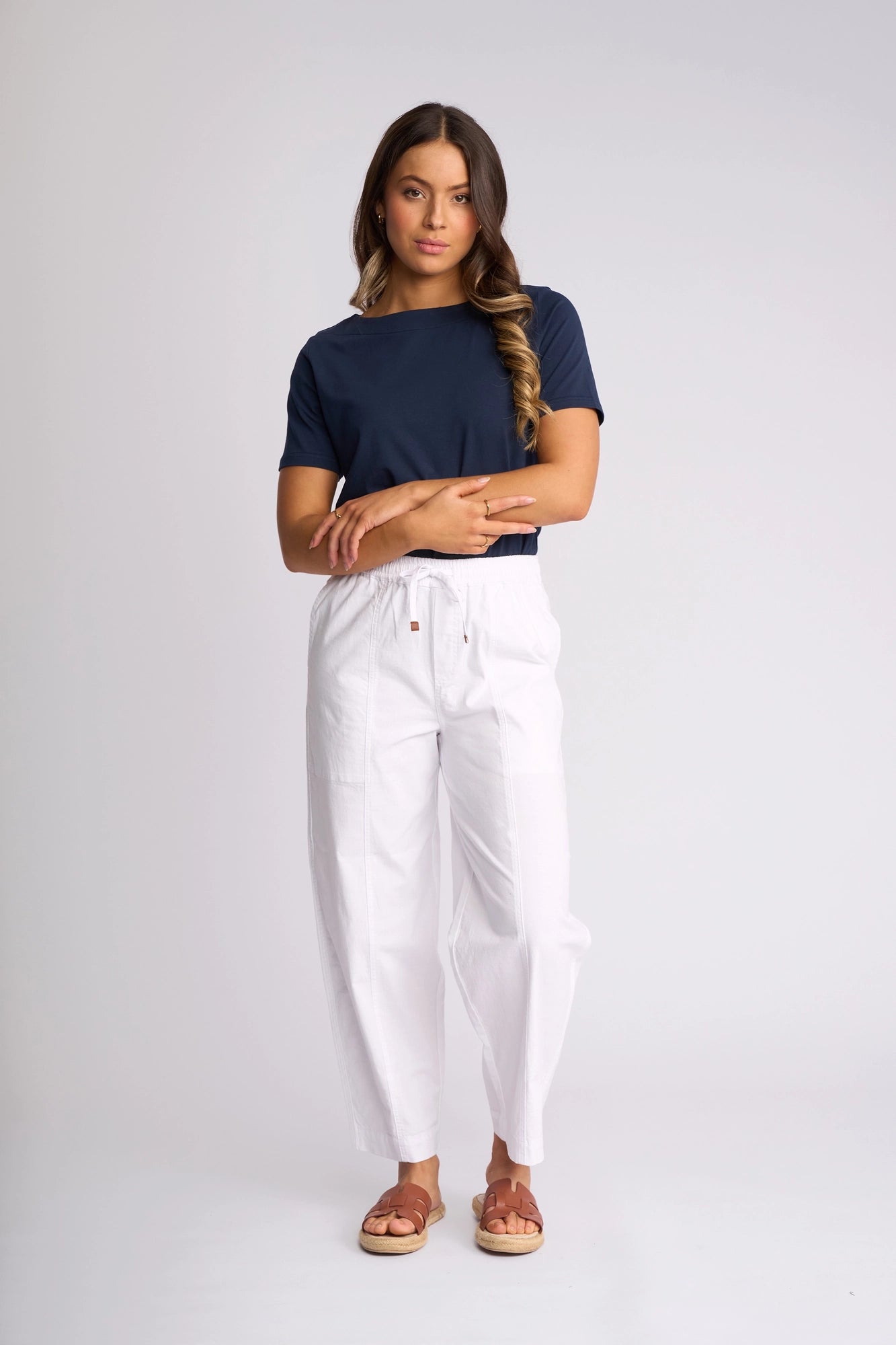 A woman stands against a plain background wearing a navy blue t-shirt, the Vassalli Dione Pant in white, and brown sandals, with her hair styled in loose waves.