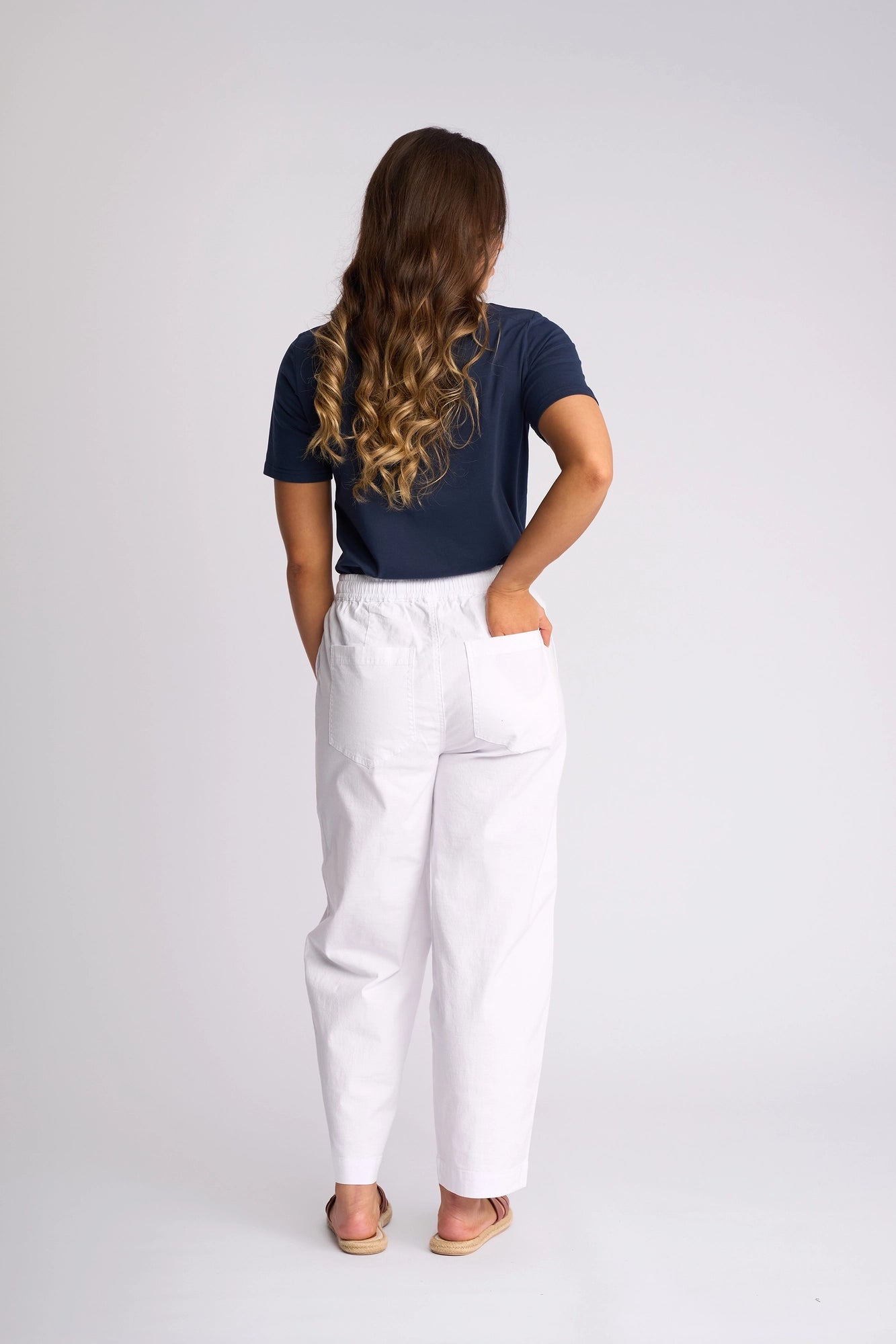 A person with long wavy brown hair, dressed in a navy blue shirt, the Vassalli Dione Pant - White, and sandals, stands facing away with one hand in their pocket against a plain light background.