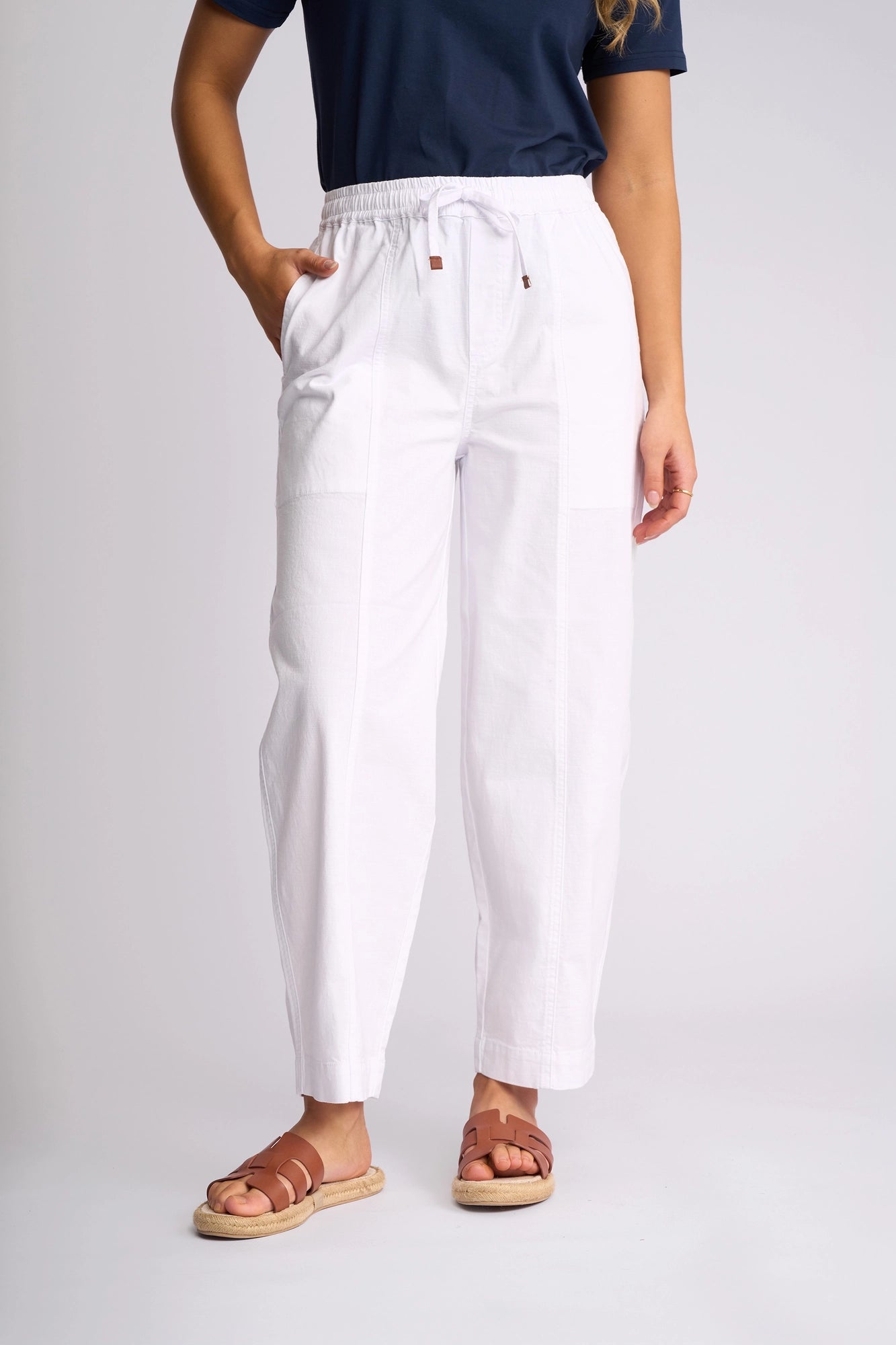 A person wearing a navy t-shirt, brown sandals, and the Vassalli Dione Pant in white stands against a plain, light background with their left hand in their pocket.