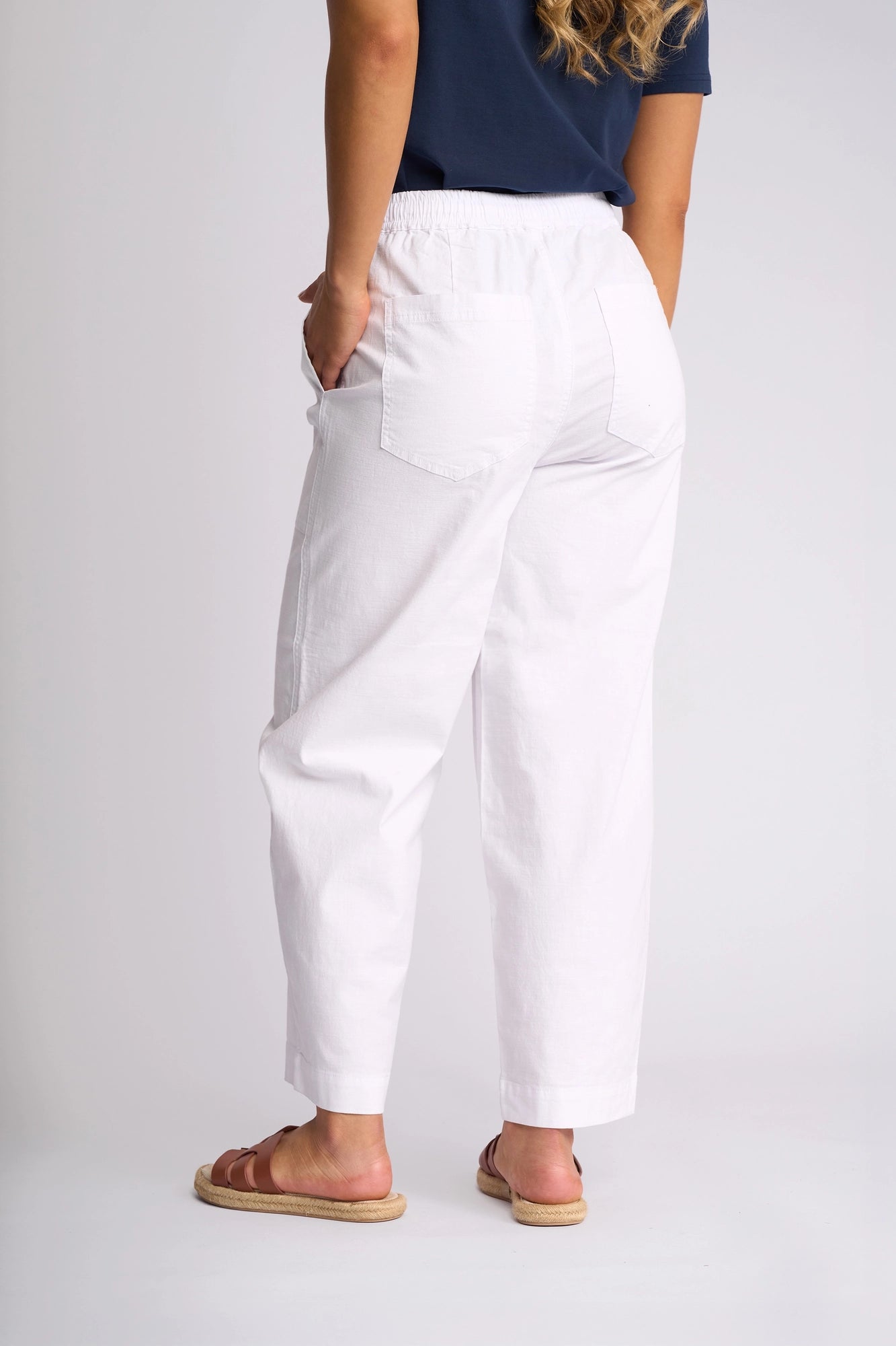 A person stands facing away, wearing the Vassalli Dione Pant in white—a stretch cotton barrel-leg style—paired with a navy shirt and brown sandals. Their right hand is tucked into their pocket, and long wavy hair rests on their shoulder. The background is plain and light.