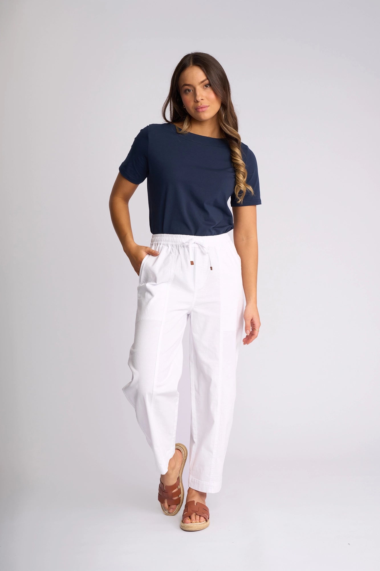 A woman stands against a plain background wearing a navy t-shirt, Vassalli Dione Pant in white—a stretch cotton barrel-leg style that grazes the ankle—and brown sandals. She has one hand in her pocket and her hair over one shoulder.