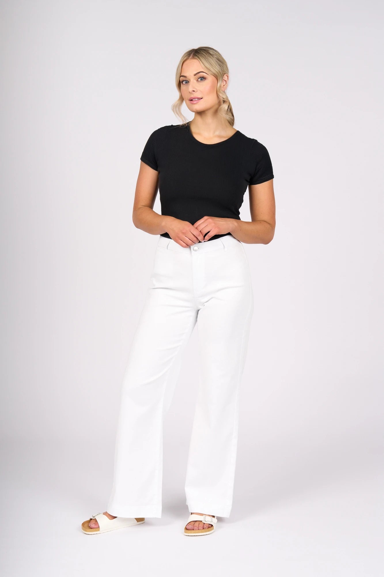 A woman stands against a plain backdrop, wearing a black short-sleeve top paired with the Vassalli Casa Jean in White Denim—a true summer staple—and white sandals. Her blonde hair falls in loose waves as she gazes slightly to the side.