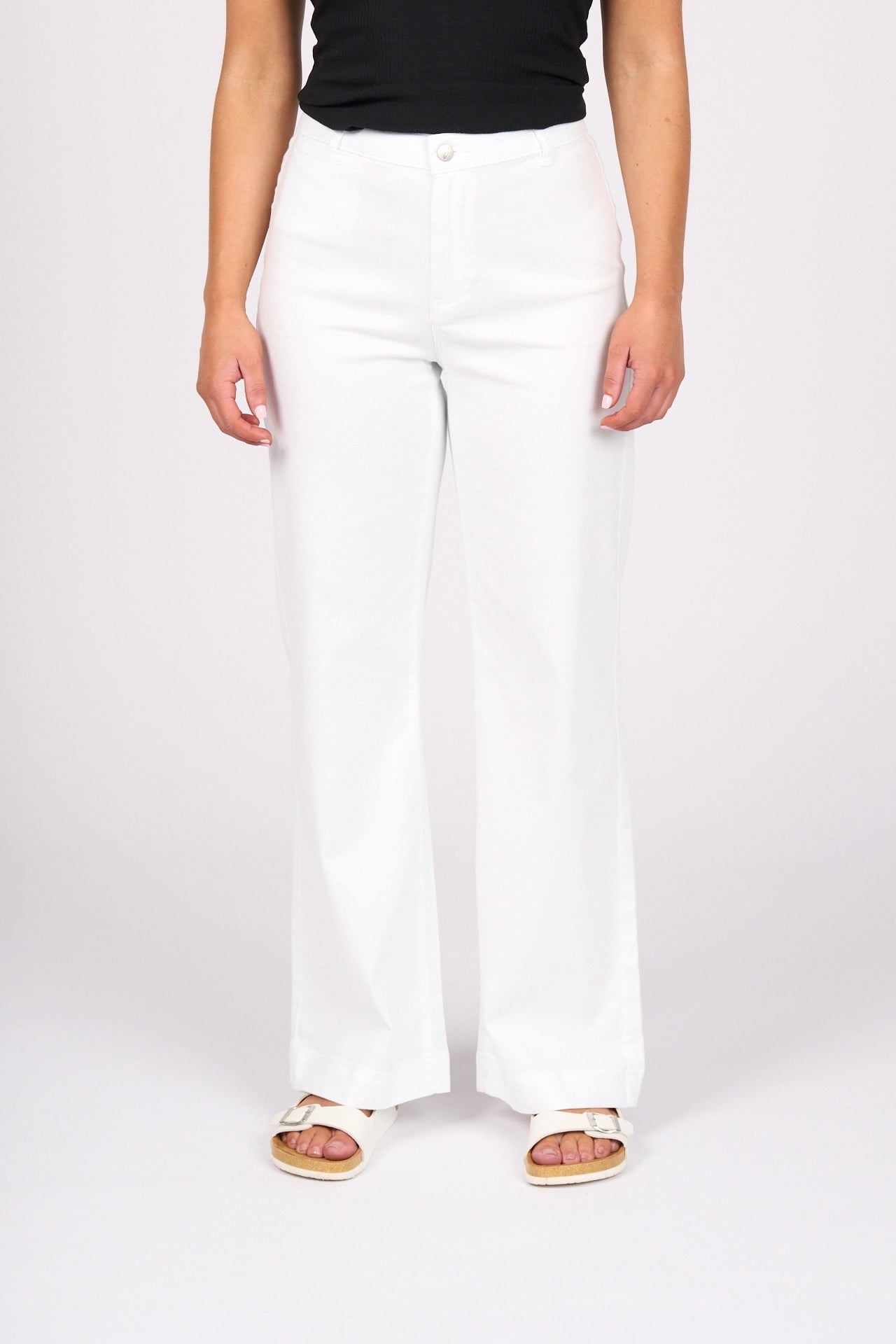 A person models the Vassalli Casa Jean in White Denim, paired with a black sleeveless top and beige sandals, standing against a plain white background. The image focuses on the outfit—showcasing this summer staple with effortless style—as their head and feet remain out of frame.