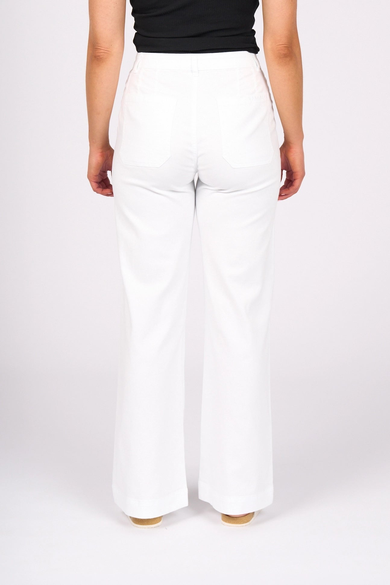 Standing with her back to the camera, she wears the Vassalli Casa Jean in white denim—this high-waisted style features classic back pockets and pairs effortlessly with a black sleeveless top for a clean, summer-ready look against a plain white background.