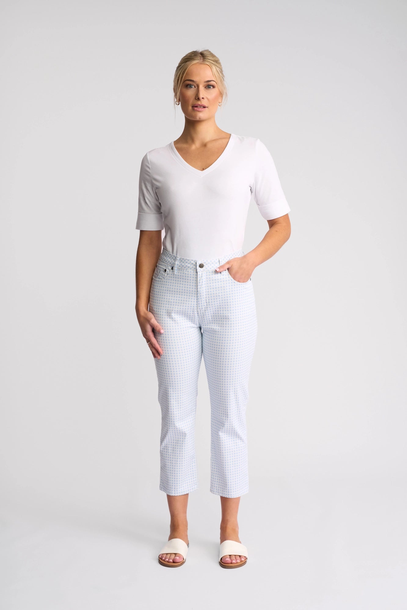 A woman stands facing forward, wearing the Vassalli Arrival Pant in Blue Gingham—slim-leg pants crafted from stretch cotton—and a white V-neck top. She completes her look with white slide sandals, one hand casually tucked in her pocket. The background is a plain light gray.