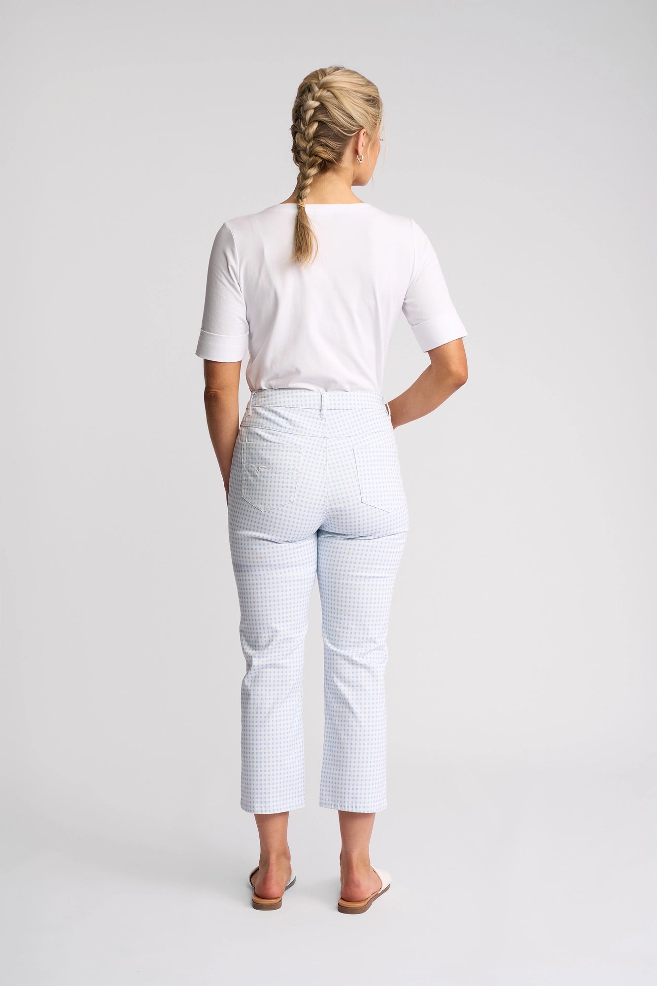 A woman with blonde hair styled in two braids, wearing a white t-shirt and the Vassalli Arrival Pant in Blue Gingham, featuring a slim-leg stretch cotton silhouette, paired with white slip-on shoes, stands facing away from the camera against a plain white background.