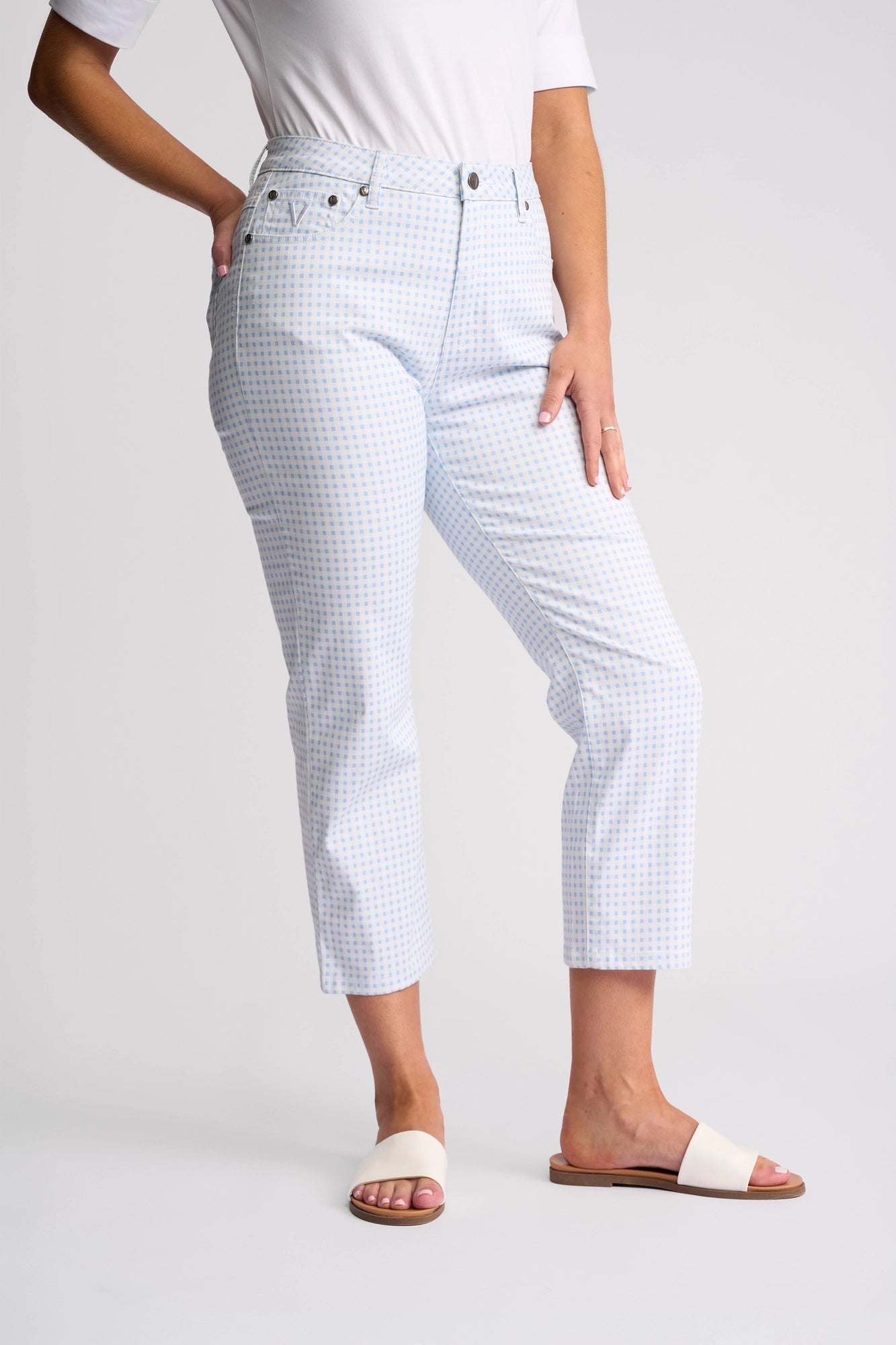 A person is posing against a plain background with one hand on their hip and the other resting on their thigh, wearing the Vassalli Arrival Pant in Blue Gingham, a crisp white shirt, and white sandals.