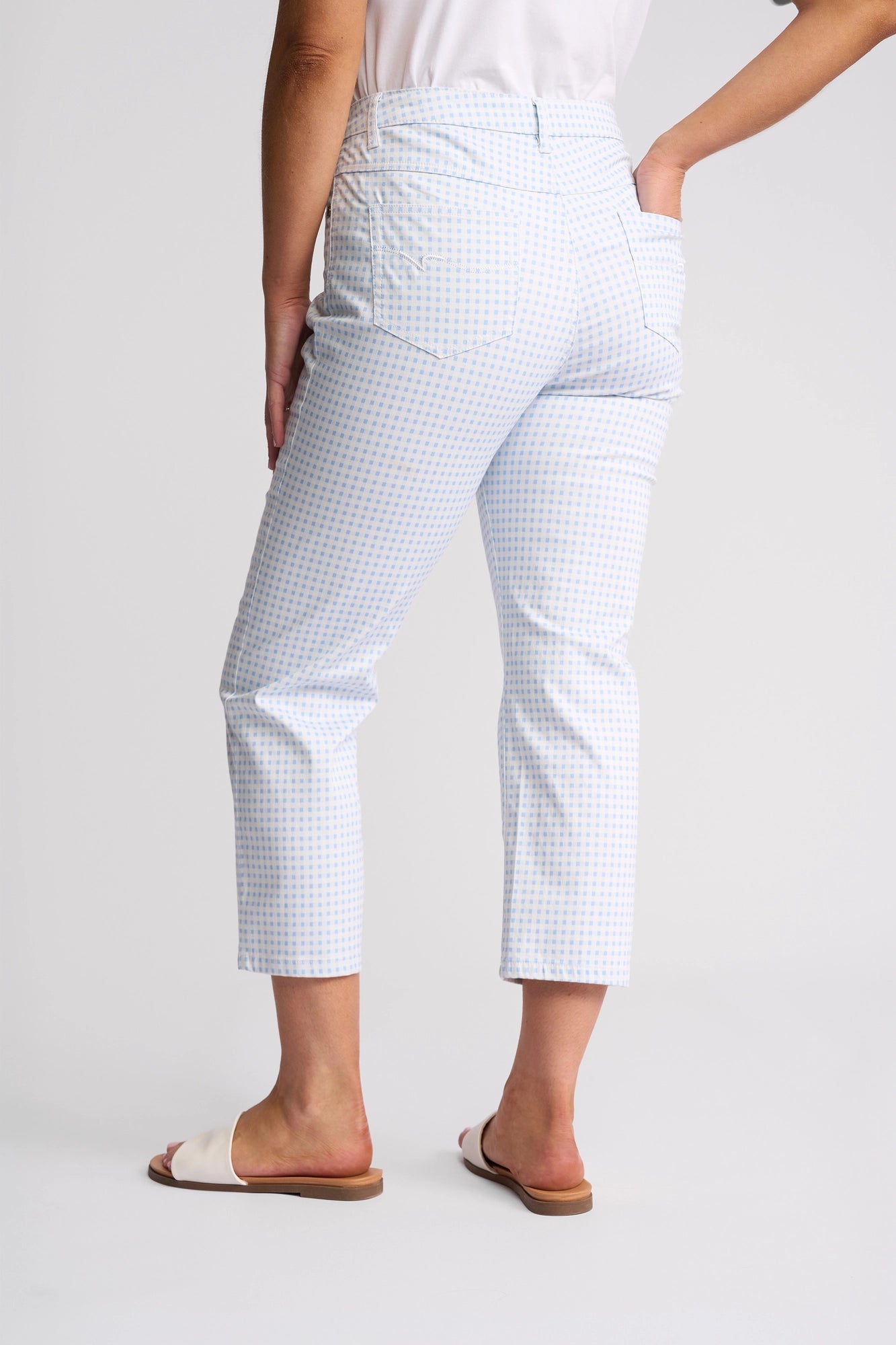 A person is seen from the back, standing against a plain light background, wearing the Vassalli Arrival Pant in Blue Gingham—a slim-leg stretch cotton style—paired with a white top and white slide sandals.