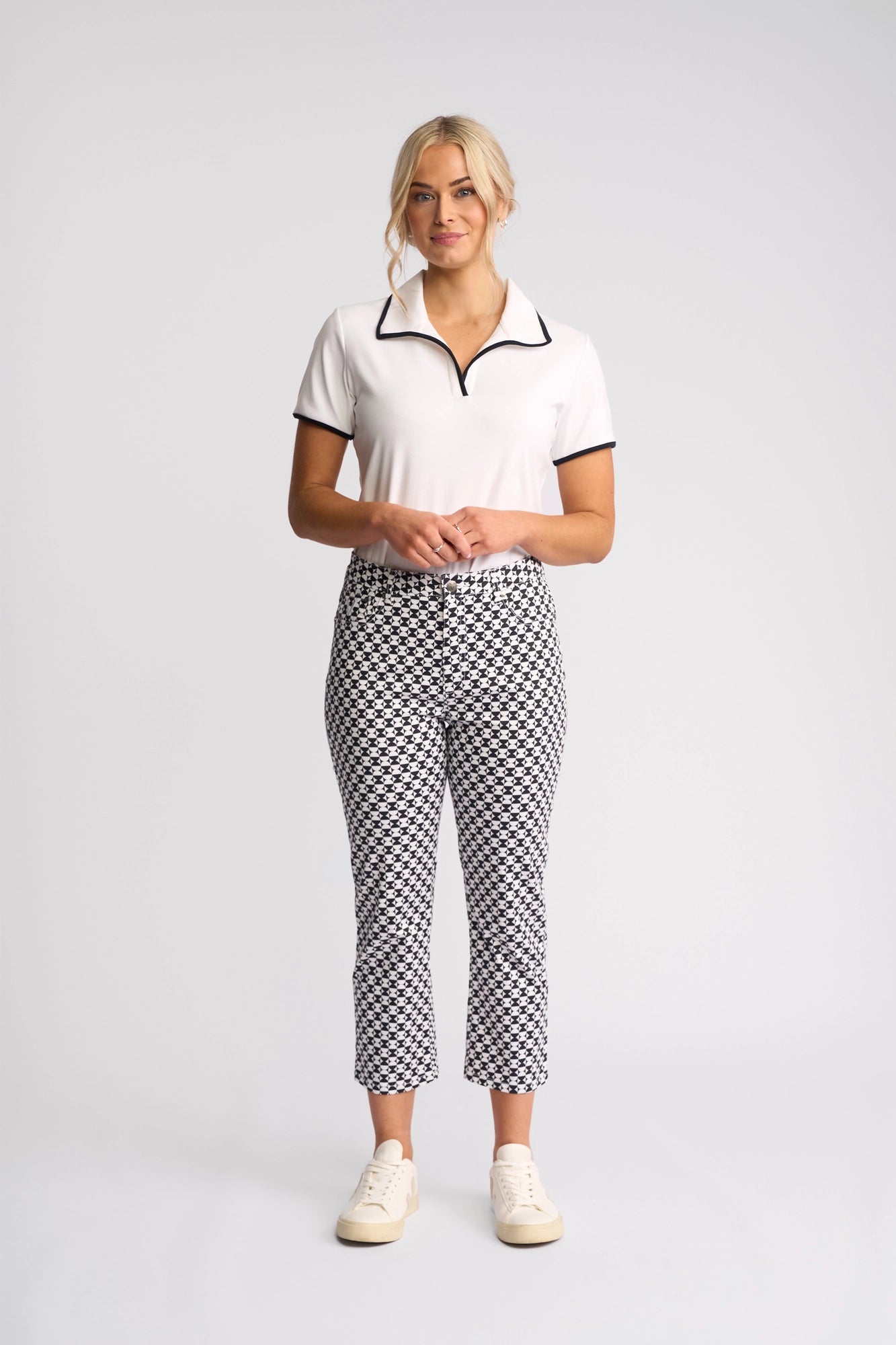 A woman stands facing forward against a plain light background, wearing the Vassalli Arrival Pant - Dixie, featuring a bold black and white geometric pattern in stretch cotton with a slim-leg fit, paired with a white collared shirt and white sneakers.