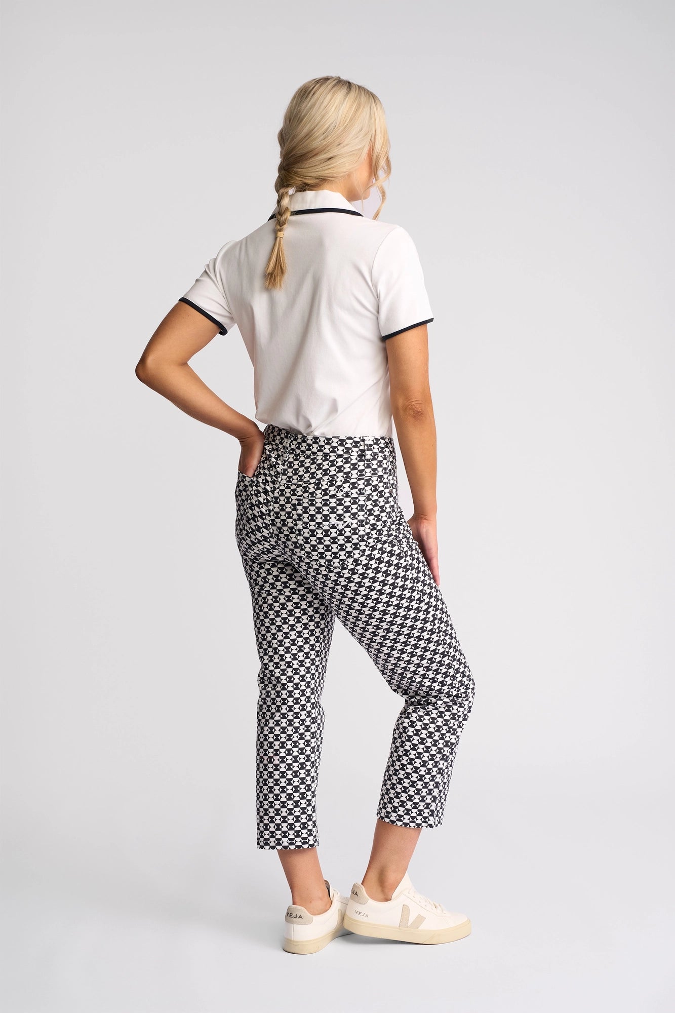A woman with blonde hair in a braid wears a white short-sleeve polo, Vassalli Arrival Pant - Dixie black and white geometric stretch cotton slim-leg pants, and white sneakers, standing with one hand on her hip and her back facing the camera.