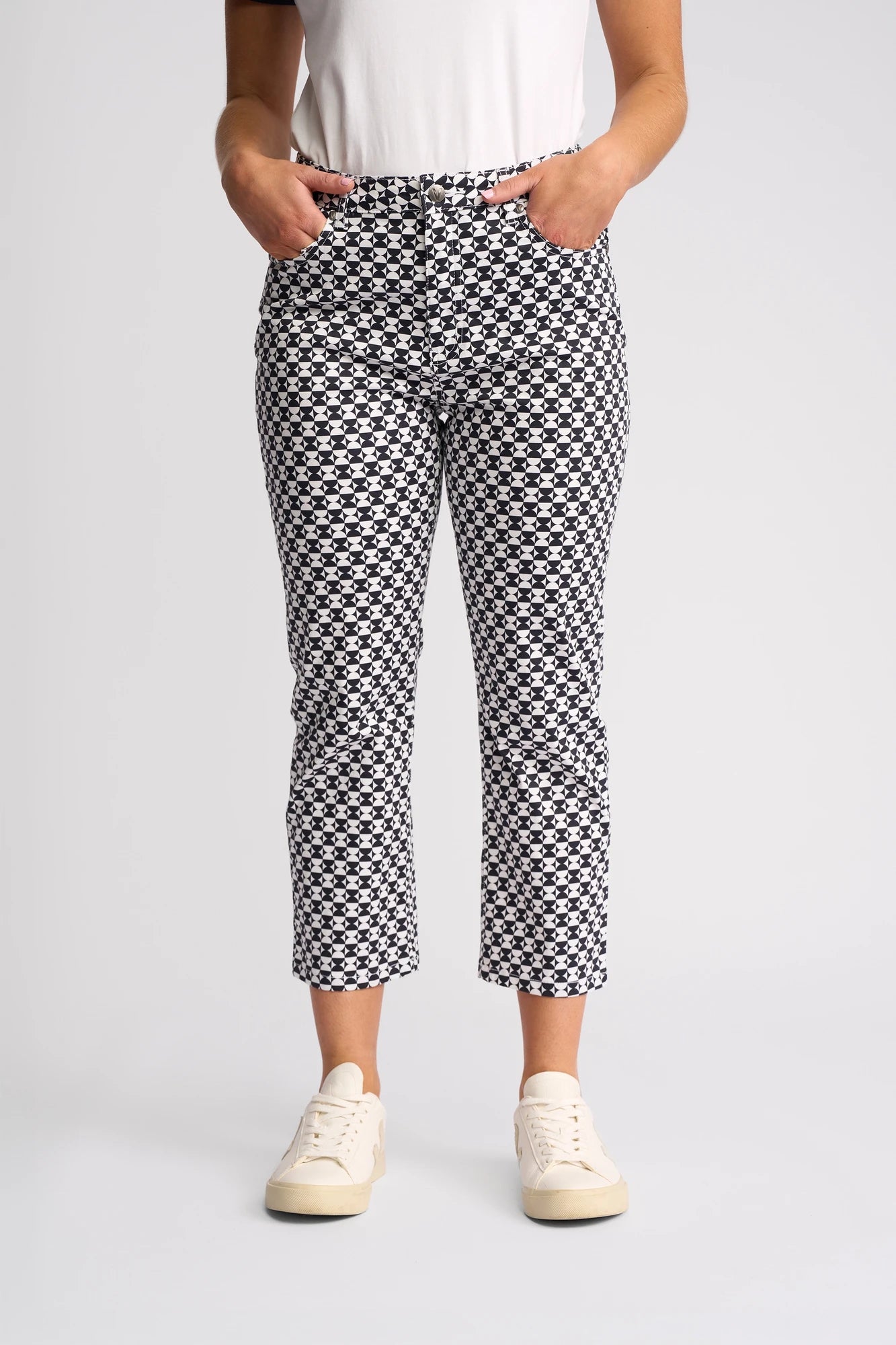 A person is wearing the Vassalli Arrival Pant - Dixie, featuring a striking black and white geometric pattern in a slim-leg style, paired with a white shirt and white sneakers, standing with hands in their pockets against a plain background.