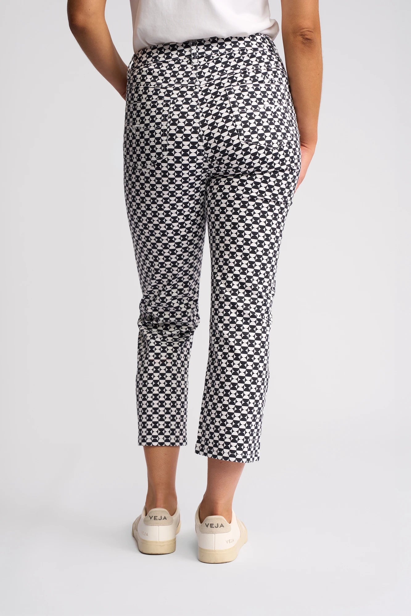 A person is standing with their back to the camera, wearing Vassalli’s Arrival Pant - Dixie: slim-leg pants in a bold black-and-white geometric check pattern, crafted from stretch cotton. They are paired with a white shirt and white Veja sneakers. The background is plain and light-colored.