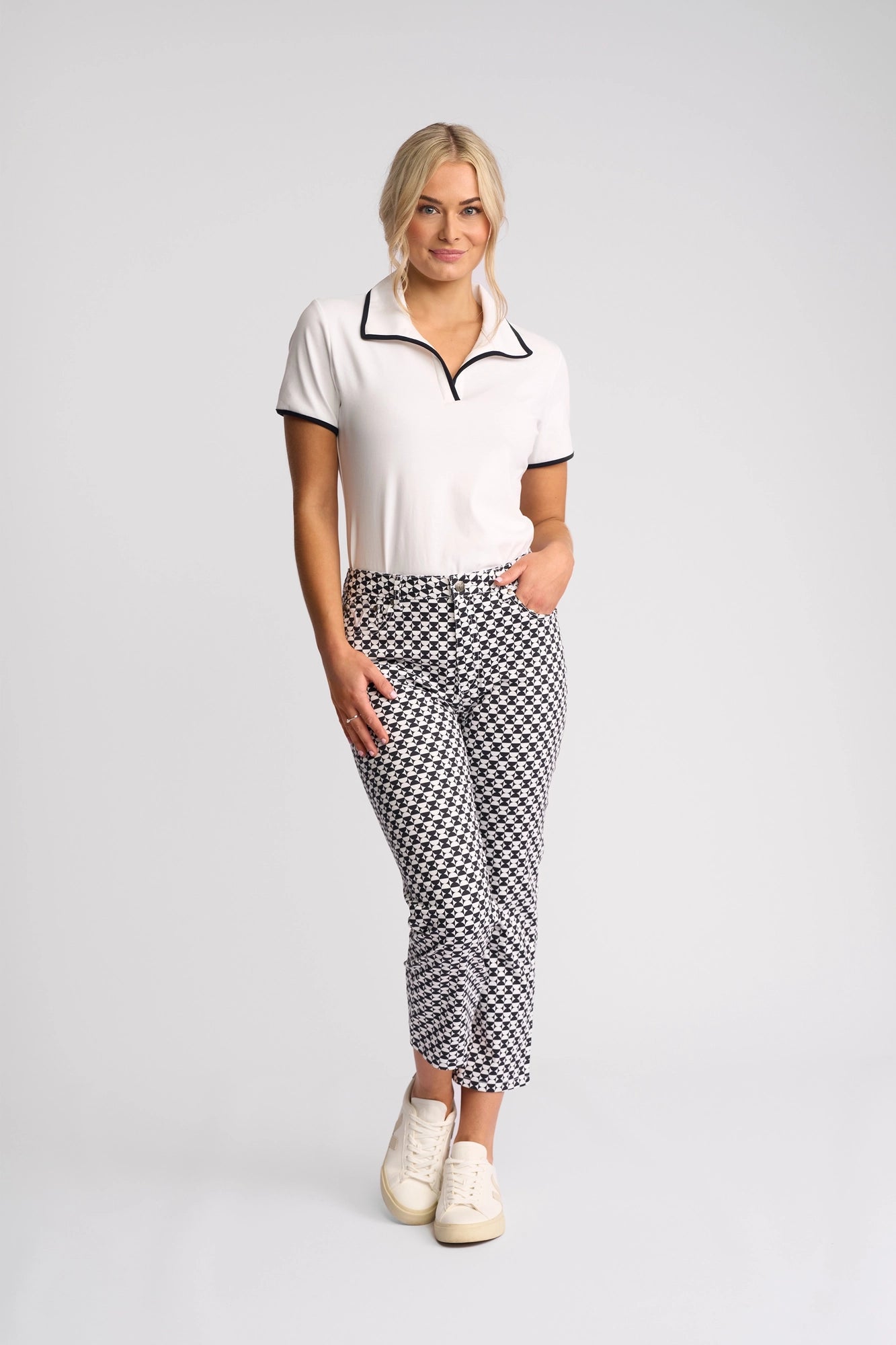 A woman stands against a plain background, wearing a white short-sleeve collared shirt, white sneakers, and the Vassalli Arrival Pant - Dixie—a pair of slim-leg pants featuring a black and white geometric pattern in stretch cotton. She poses with one hand in her pocket and smiles slightly.