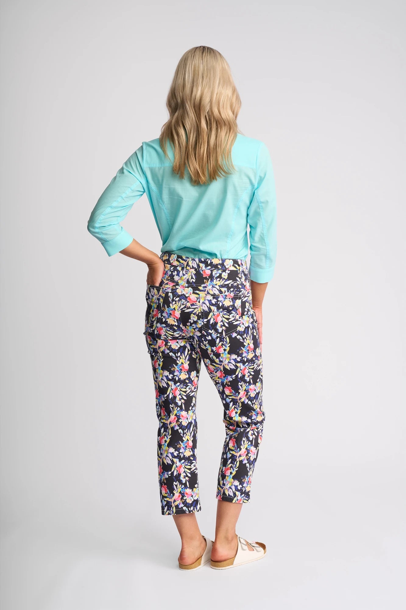 A woman with wavy blonde hair stands facing away against a plain, light background, wearing a light blue long-sleeve shirt paired with the Vassalli Arrival Pant - Lani. The slim-leg pants feature a floral design and are crafted from comfortable stretch cotton.
