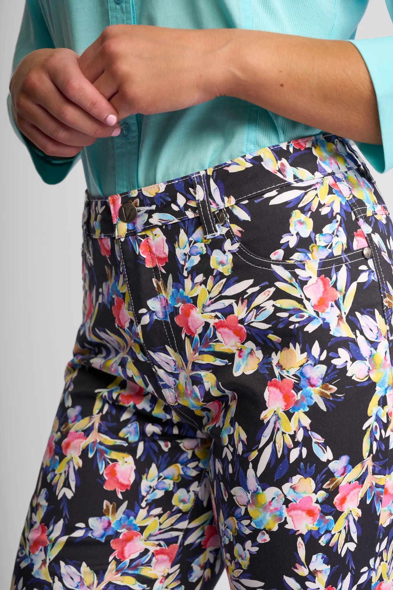 A person wearing a light teal button-up shirt and high-waisted, slim-leg Vassalli Arrival Pant - Lani in black with colorful floral print. The Arrival Pant by Vassalli features stretch cotton for comfort. Only the torso and upper legs are visible.
