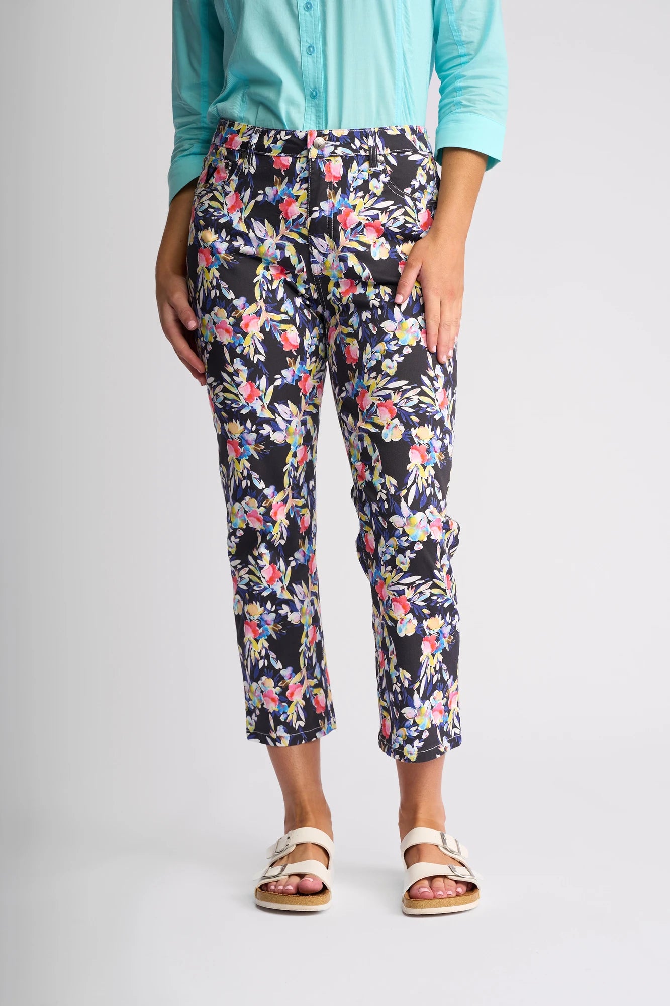 A person, visible only from the waist down, stands against a plain light background wearing a turquoise long-sleeve shirt, vibrant floral slim-leg stretch cotton cropped pants—the Vassalli Arrival Pant - Lani—and white sandals.