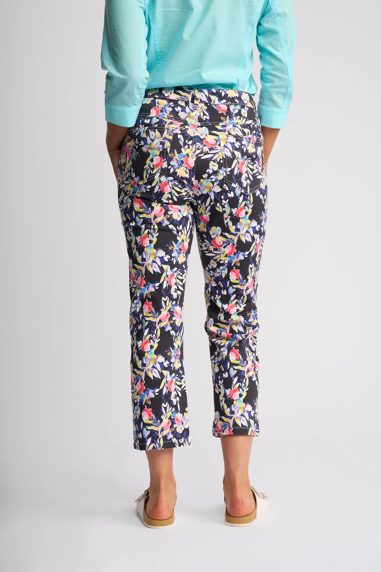 A person stands facing away, barefoot on a light background, wearing a light blue long-sleeve shirt paired with the Vassalli Arrival Pant - Lani. These ankle-length, slim-leg pants feature vibrant floral patterns and are crafted from stretch cotton for both comfort and style.