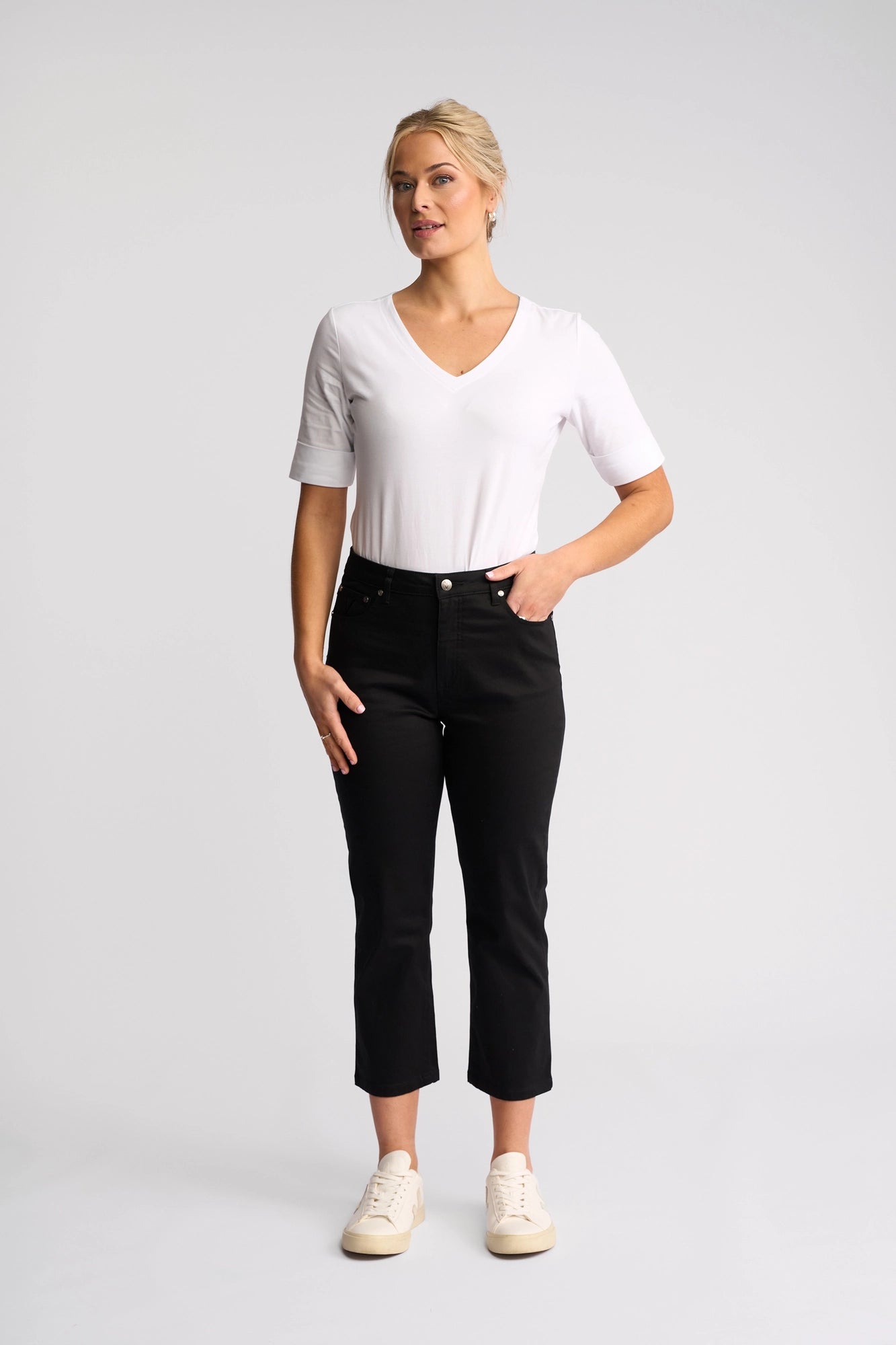 A woman stands facing forward with one hand in her pocket against a plain light background, wearing a white V-neck shirt, white sneakers, and the Vassalli Arrival Pant in Black.