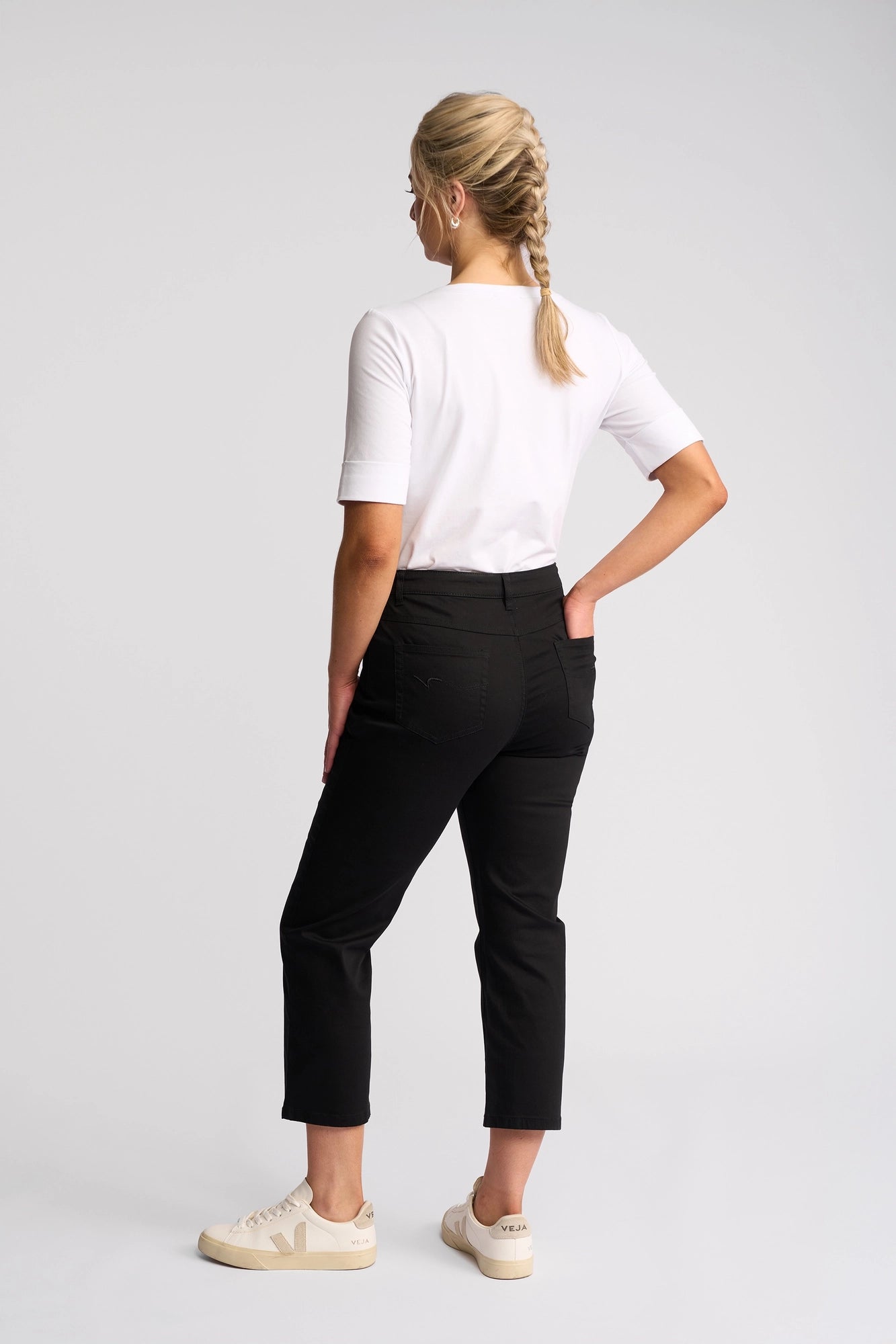 A person with blonde hair in a braid, dressed in a white shirt, Vassalli Arrival Pant in black, and white sneakers stands facing away against a plain white background.