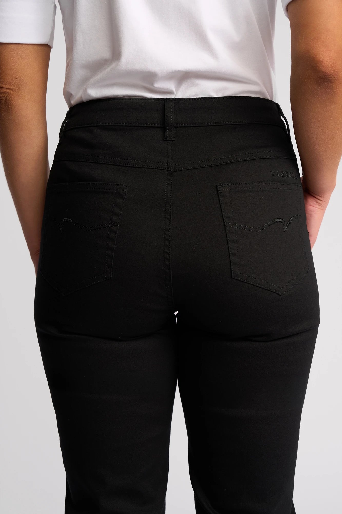 A person is shown from the back, focusing on the waist and hips, wearing the Vassalli Arrival Pant - Black. This slim-leg pant is crafted from stretch cotton and features two back pockets with subtle stitching.