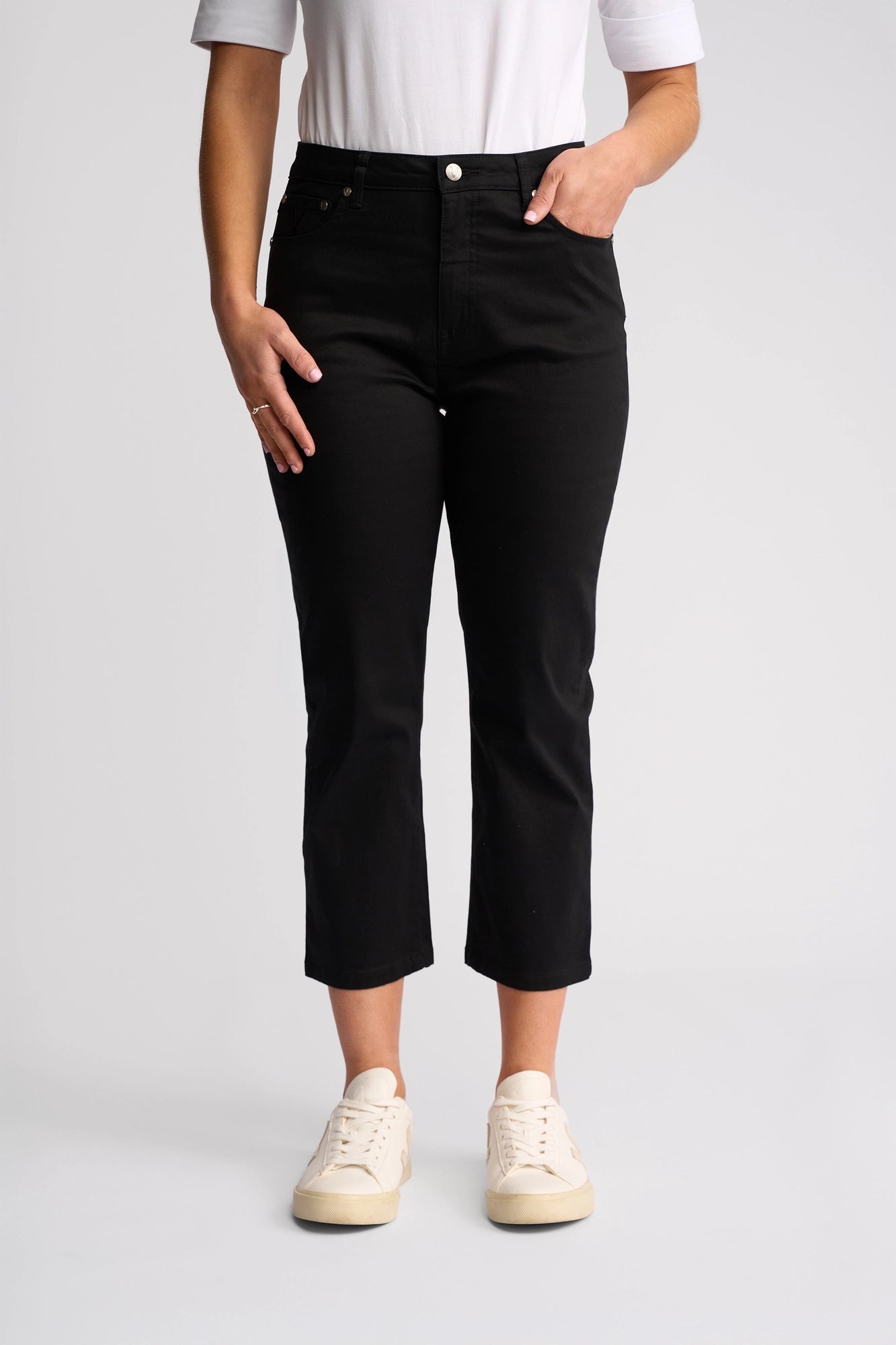 A person wearing a white shirt, the Vassalli Arrival Pant - Black in a slim-leg cut crafted from stretch cotton, and white sneakers stands against a plain light background with one hand in their pocket and the other resting by their side.