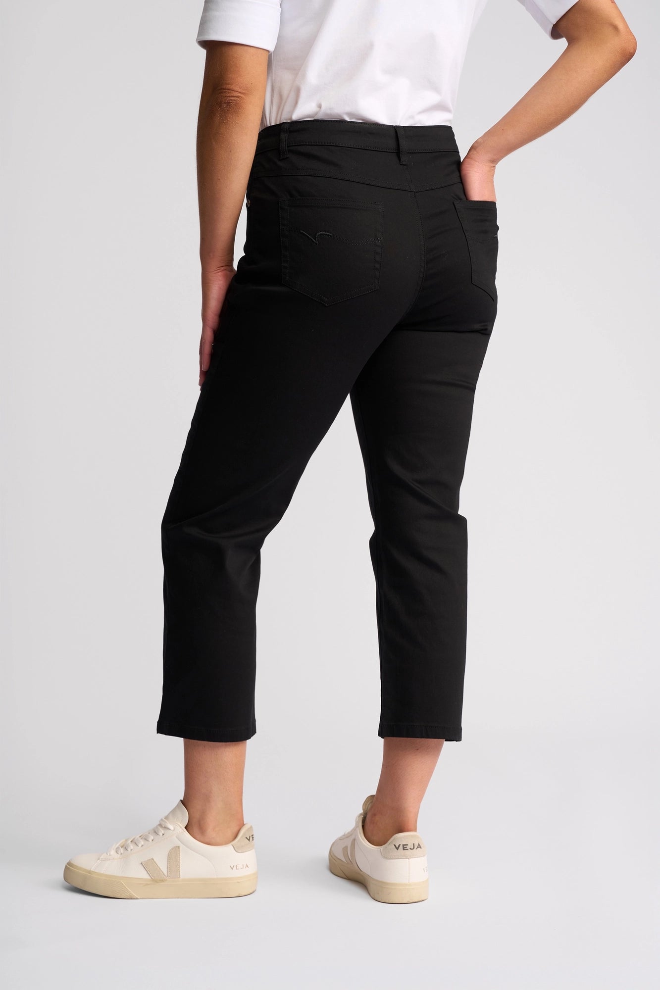 A person seen from behind wearing the Vassalli Arrival Pant in black—cropped slim-leg pants crafted from stretch cotton—paired with a white t-shirt and white Veja sneakers, with one hand in their back pocket.