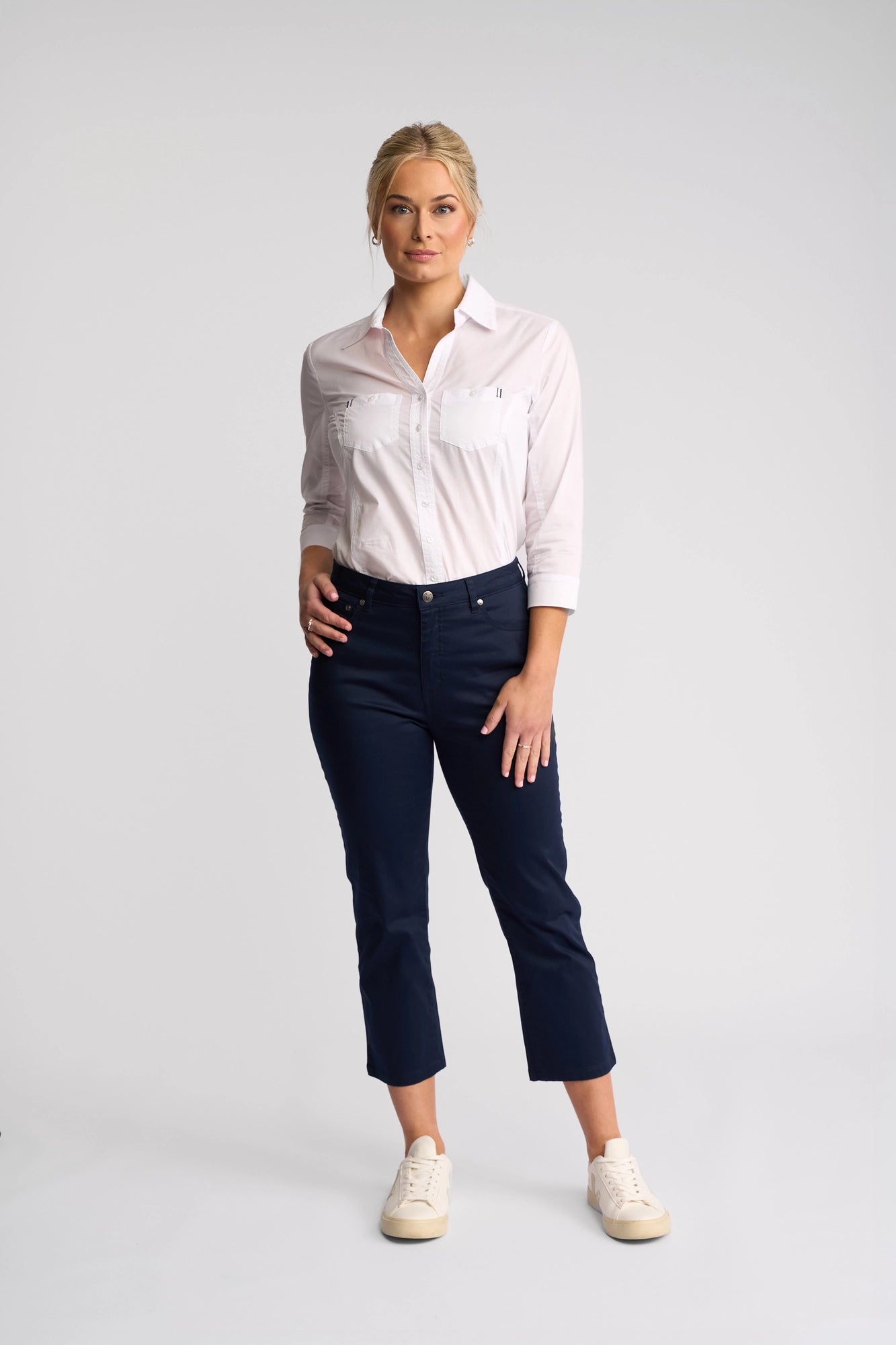 A woman stands facing forward, wearing a light pink button-up shirt, Vassalli Arrival Pant in navy crafted from stretch cotton, and white sneakers against a plain light background.