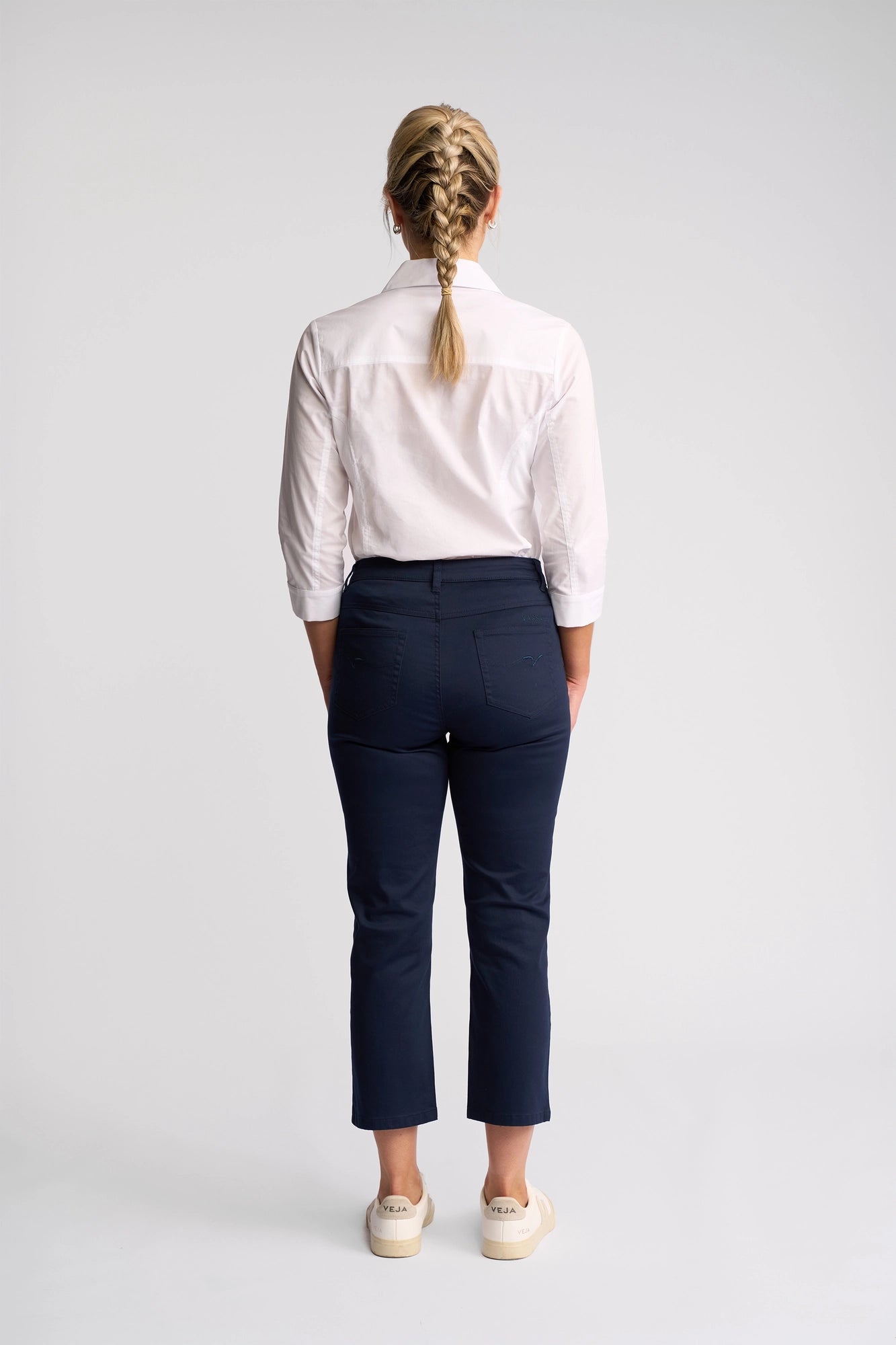 A person with blonde hair in a braid is standing facing away, wearing a white button-up shirt, Vassalli Arrival Pant in navy, and beige shoes against a plain light background.
