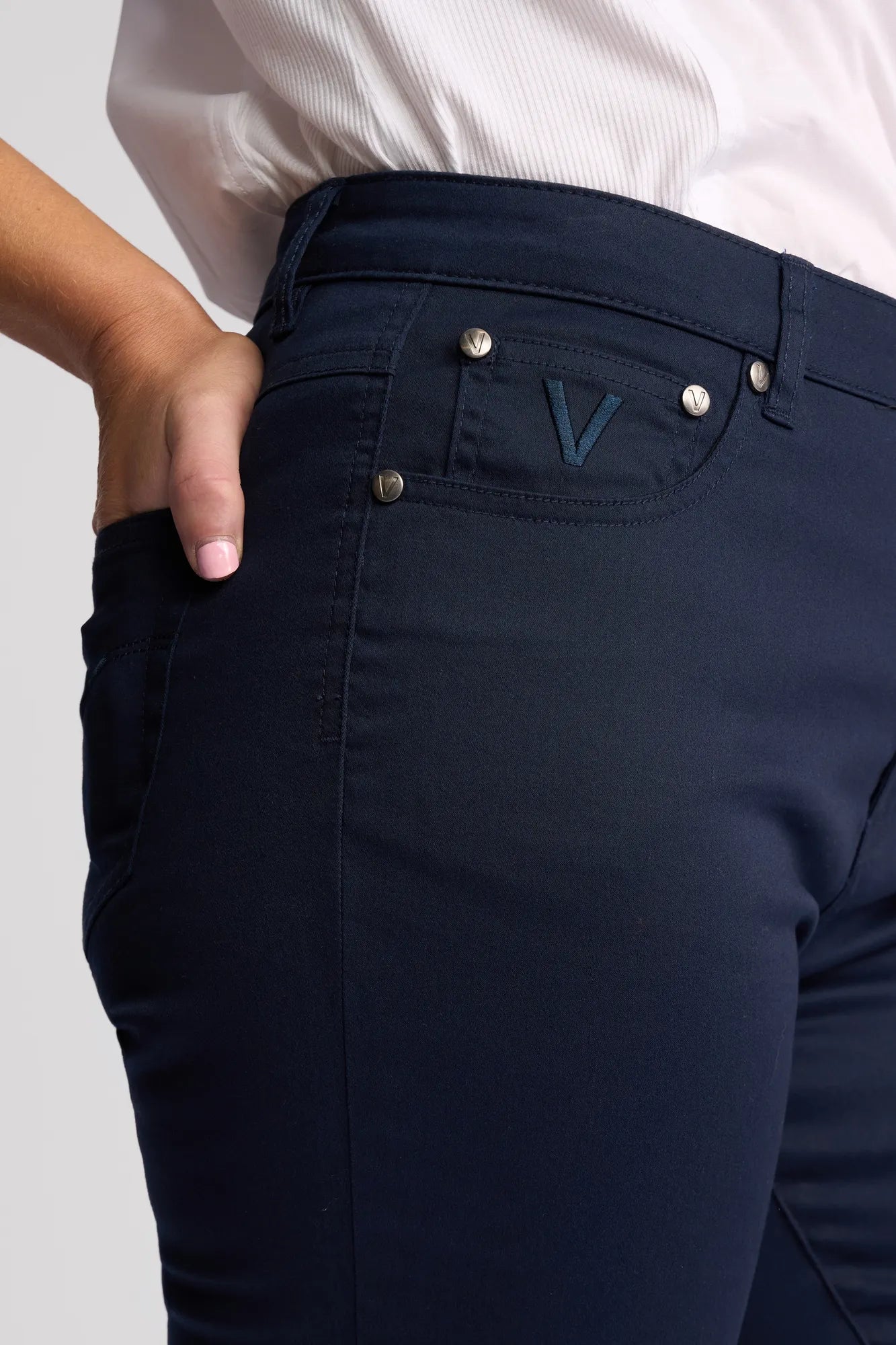 Dressed in the Vassalli Arrival Pant - Navy, featuring a signature V logo on the pocket, a person places their left hand into the front pocket. The slim-leg, stretch-cotton pant is styled with a tucked-in white shirt for a sleek appearance.