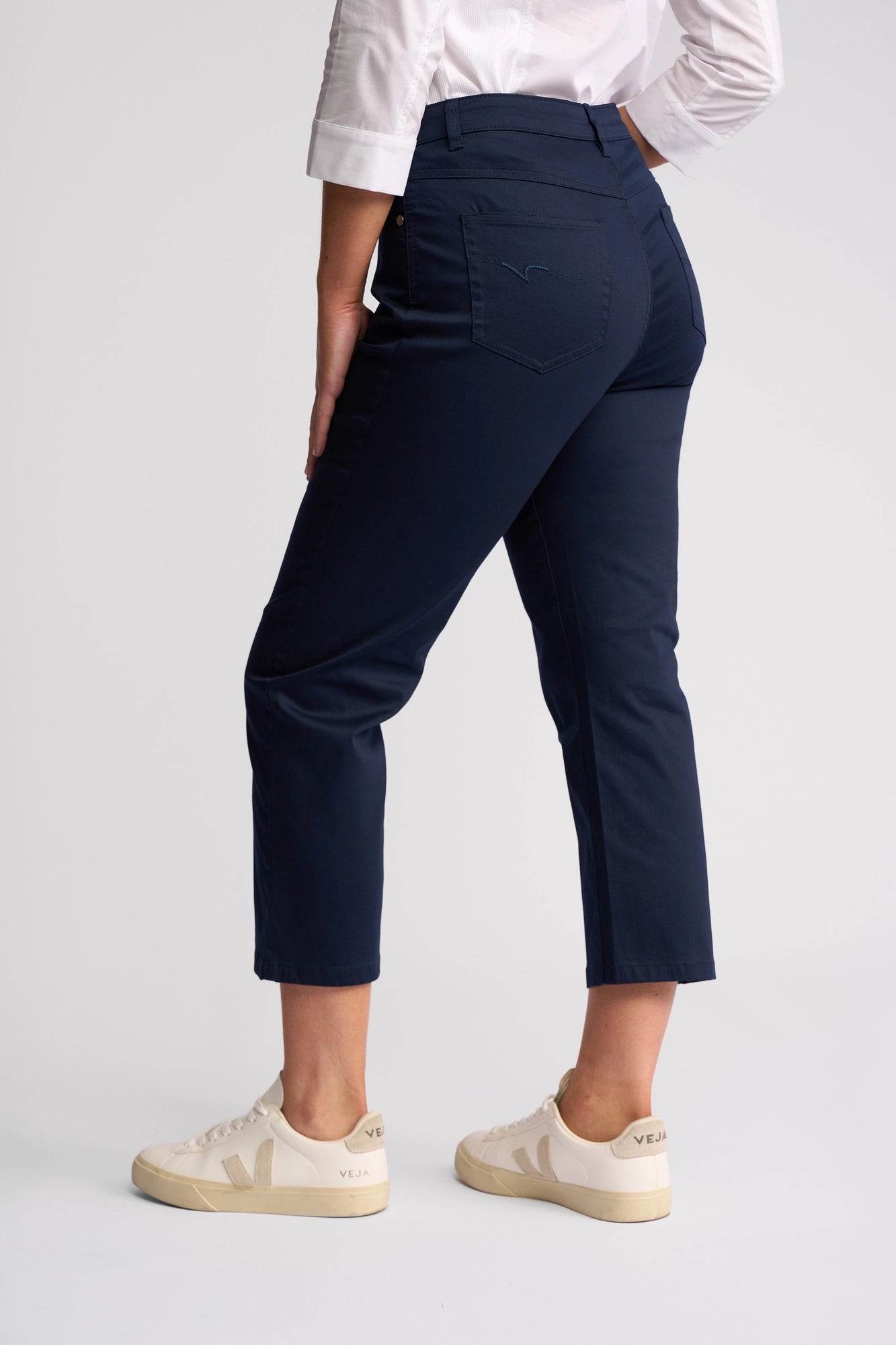 A person wearing the Vassalli Arrival Pant - Navy, paired with a white shirt featuring rolled sleeves and white sneakers, stands against a plain light background, shown from the back and slightly turned to the side. The slim-leg Arrival Pant by Vassalli is made from stretch cotton for comfortable, effortless style.