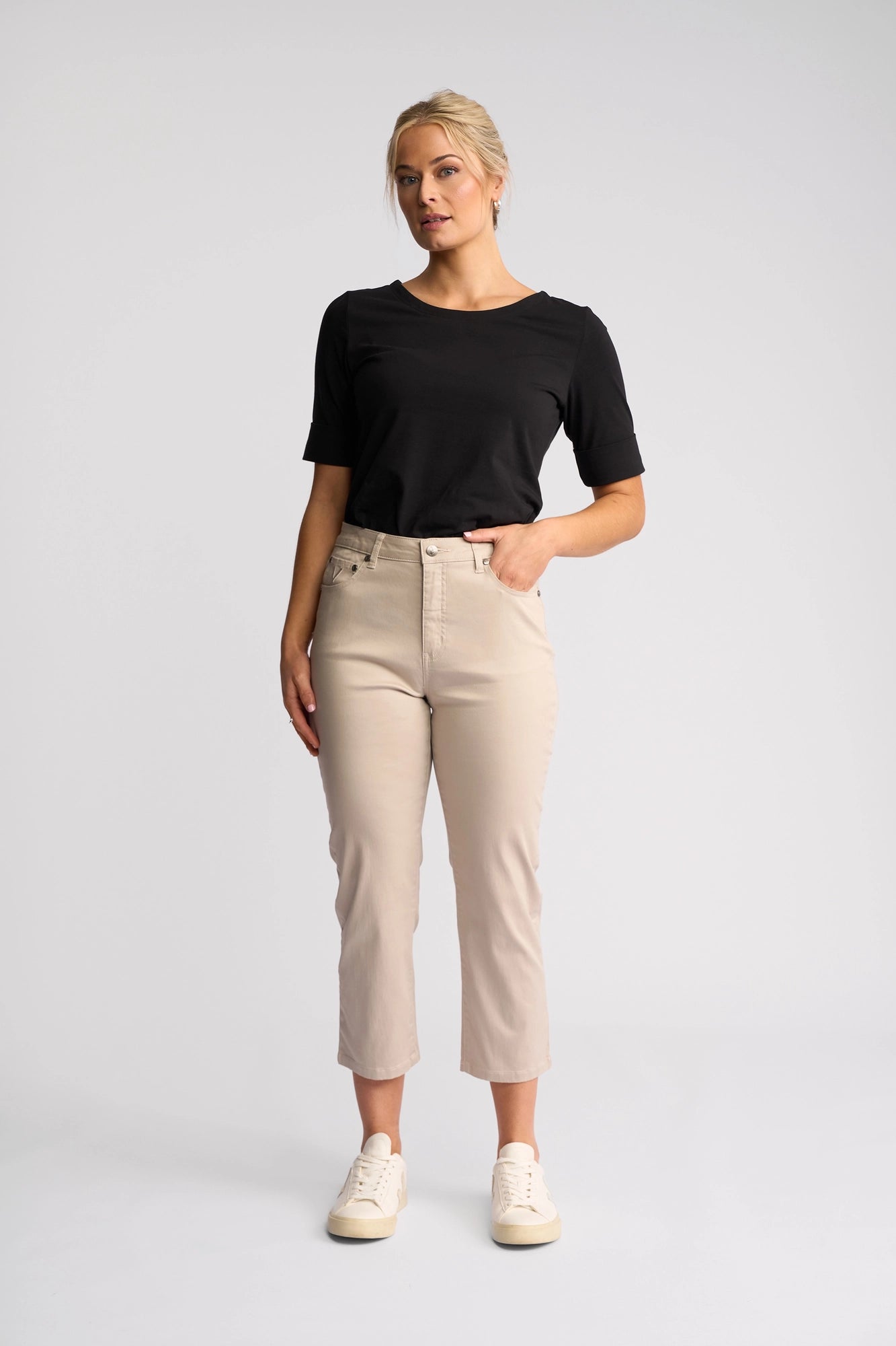 A woman stands facing forward, wearing a black short-sleeve top paired with the Vassalli Arrival Pant in Pumice—slim-leg beige pants crafted from stretch cotton—and light sneakers. Her left hand rests in her pocket as she poses against a plain light gray background.