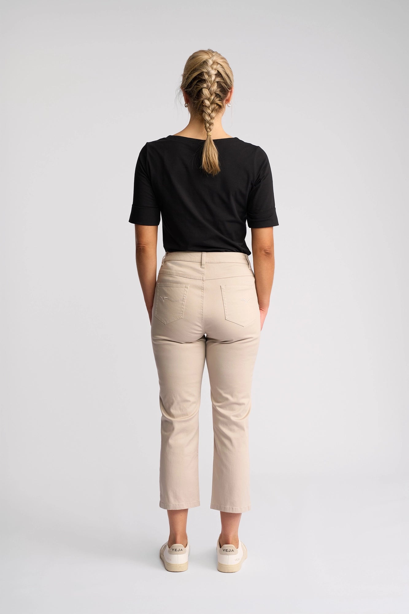 A woman with blonde hair in a braid stands facing away, wearing a black short-sleeve shirt, white sneakers, and the Vassalli Arrival Pant in Pumice—a slim-leg style crafted from stretch cotton—against a plain light background.