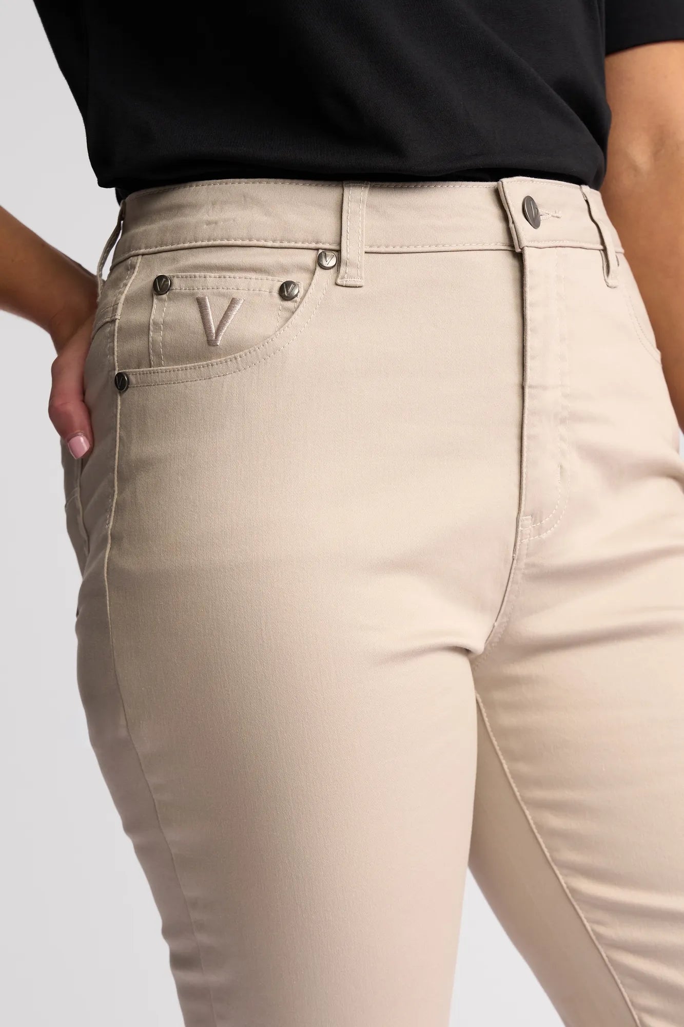 A person wearing high-waisted Vassalli Arrival Pant in Pumice and a black shirt, with one hand resting on their hip. The slim-leg pants, crafted from stretch cotton, feature front pockets and a stitched V detail near the pocket.