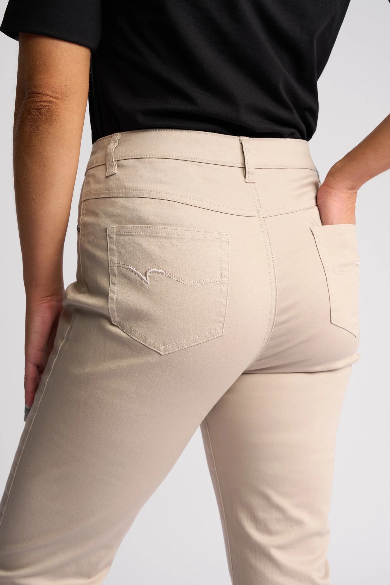 A person is shown from the waist down wearing the Vassalli Arrival Pant in Pumice, a stretch cotton slim-leg style, paired with a black shirt and posing with one hand on their hip. The pants feature two back pockets with subtle stitching.