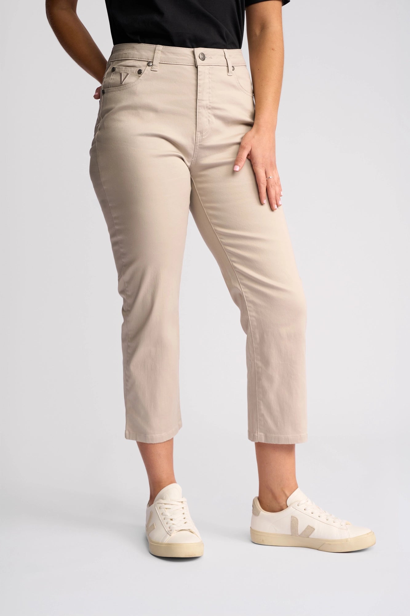 A person wearing Vassalli’s Arrival Pant in Pumice—a slim-leg pant made from stretch cotton—paired with a black shirt and white sneakers, stands against a plain white background with one hand resting on their thigh.